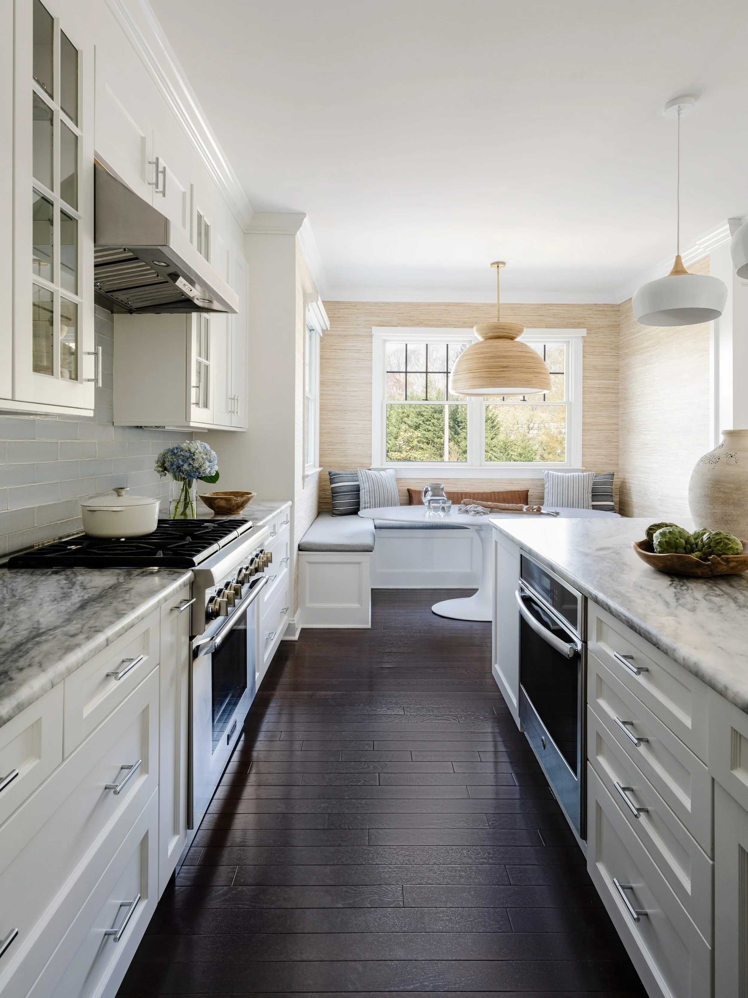 Galley kitchen with white shaker cabinetry, marble countertops, glass-front upper cabinets, stainless steel range and range hood, dark wood flooring, built-in breakfast nook with blue cushion and striped pillows beneath window with natural grasscloth