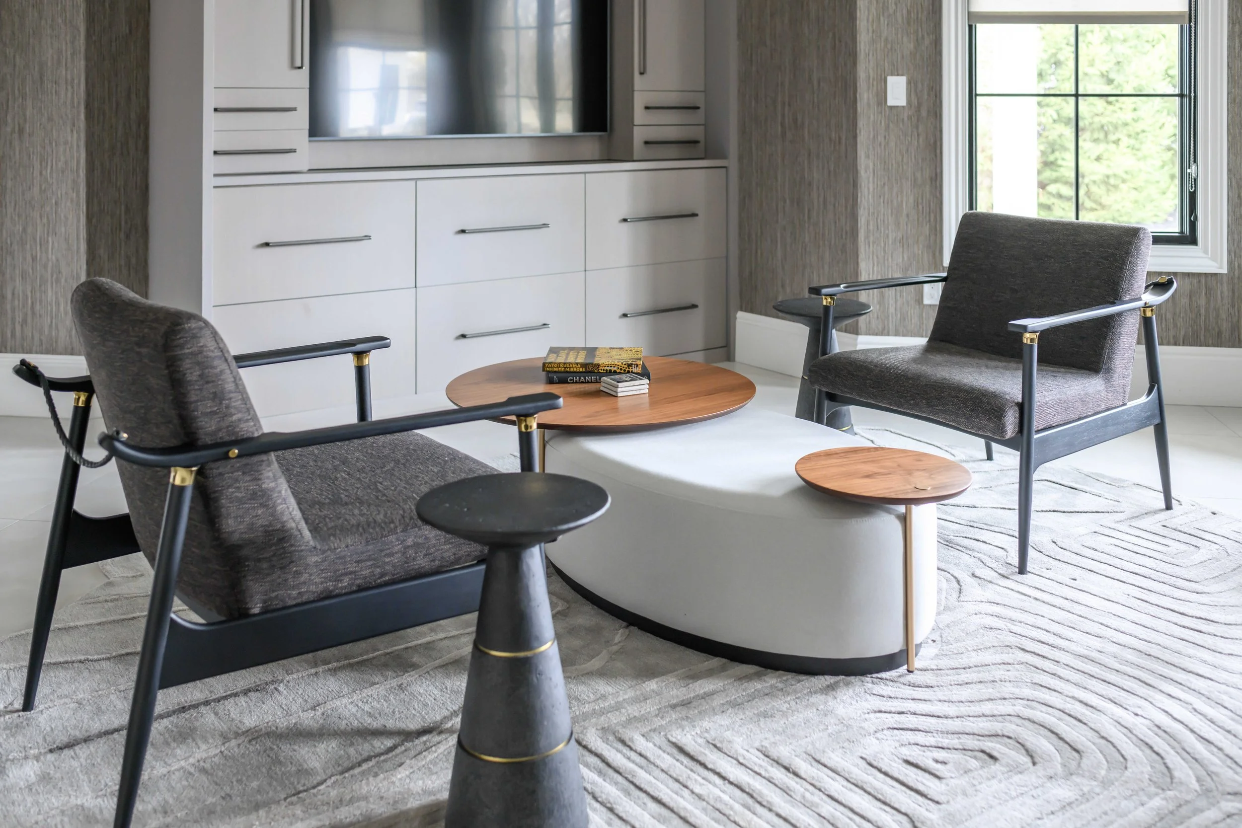 Sophisticated seating area with two gray upholstered mid-century chairs with black frames and brass accents, tiered round coffee tables in wood and black stone, white built-in entertainment center, and textured gray grasscloth wallcovering.