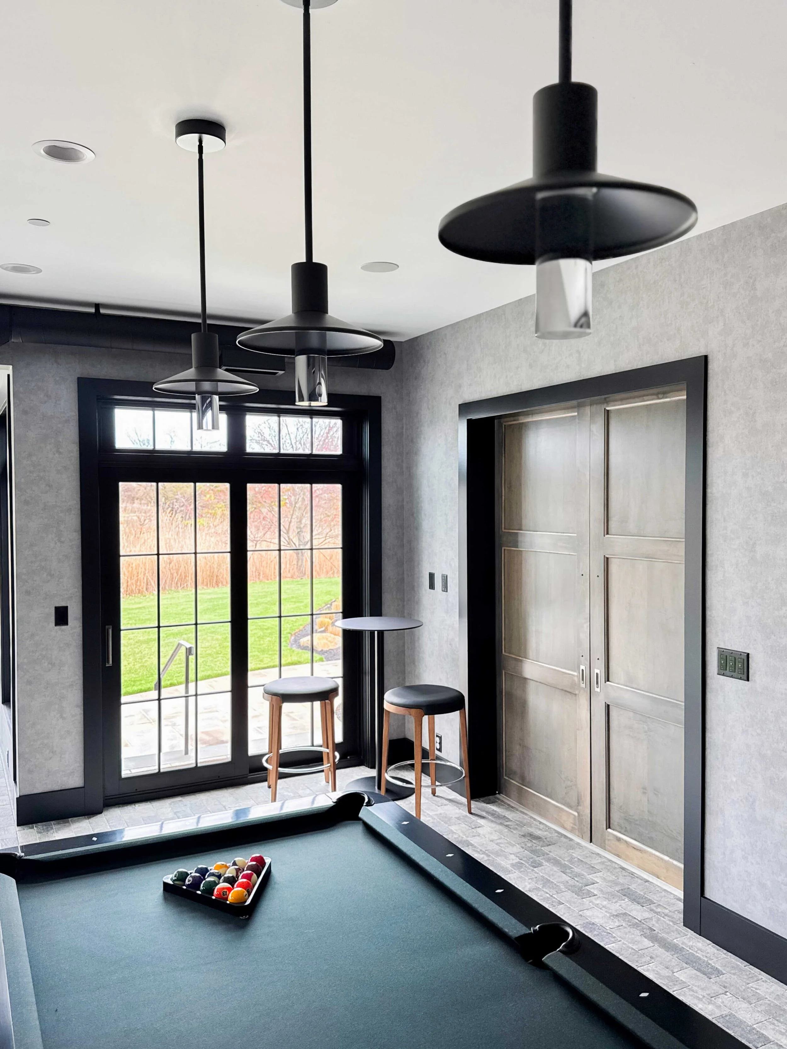 Modern game room with black-framed glass door opening to lawn, pool table with racked balls, black disc pendant lights, two wood and black leather bar stools at high-top table, and residential elevator with paneled doors.