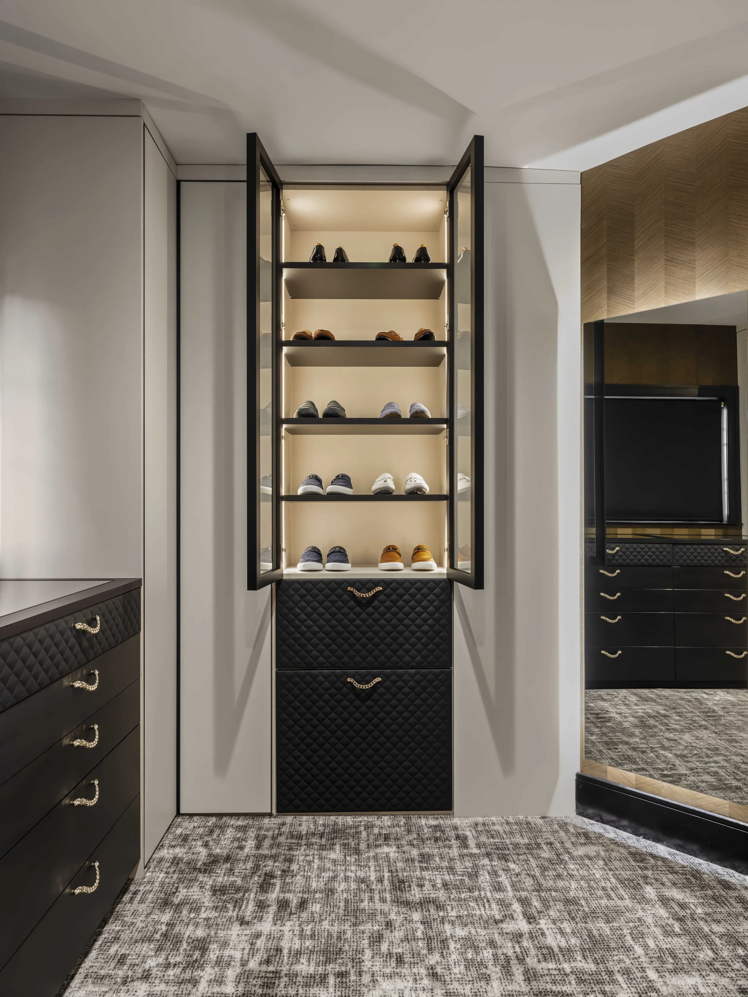 Detailed view of an open glass-front shoe cabinet showcasing designer footwear, featuring black quilted drawer bases and a grey textured rug.
