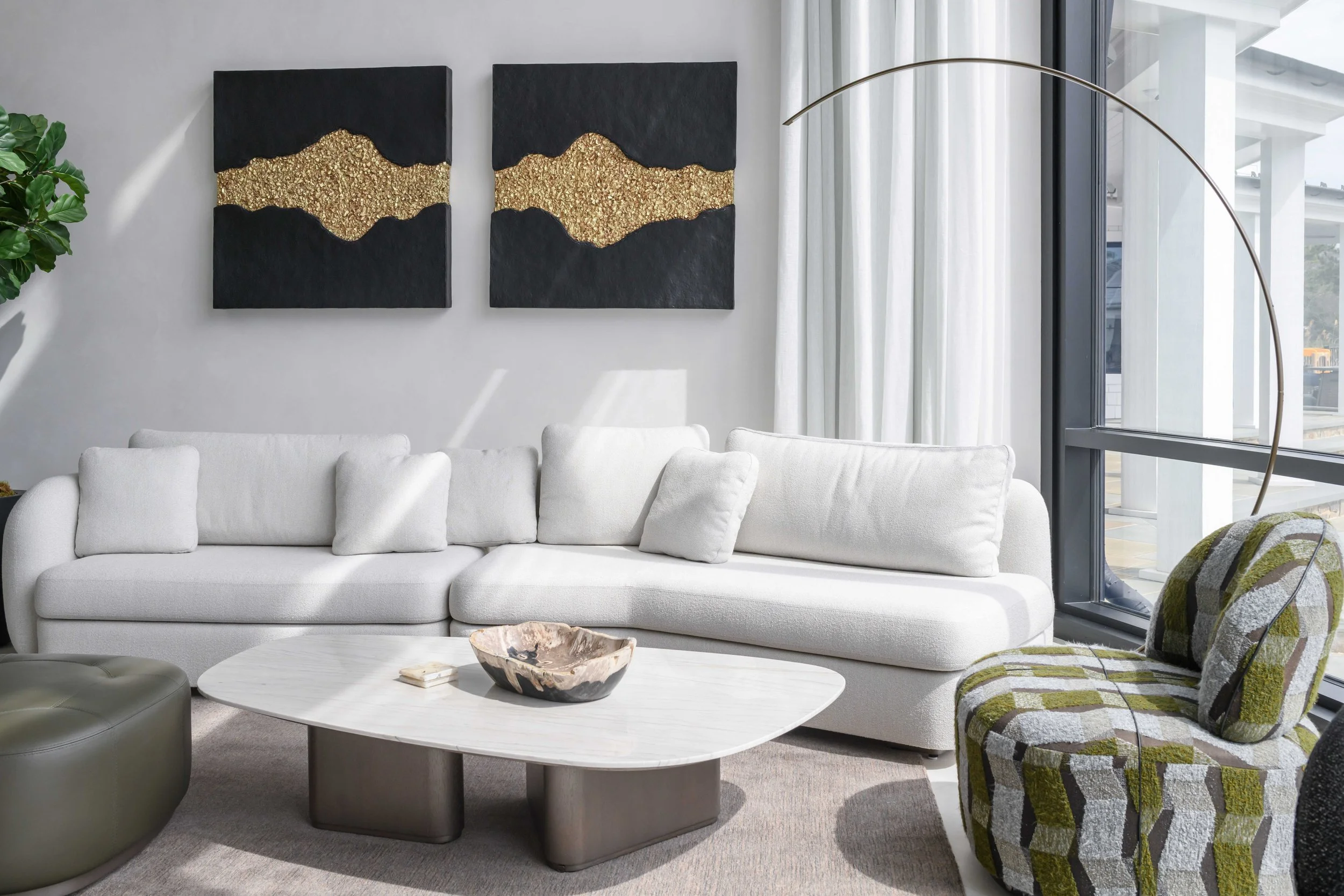 Modern living room with light gray sectional sofa, black and gold abstract wave artwork, sculptural white coffee table, arching floor lamp, and patterned green accent chair near large windows.