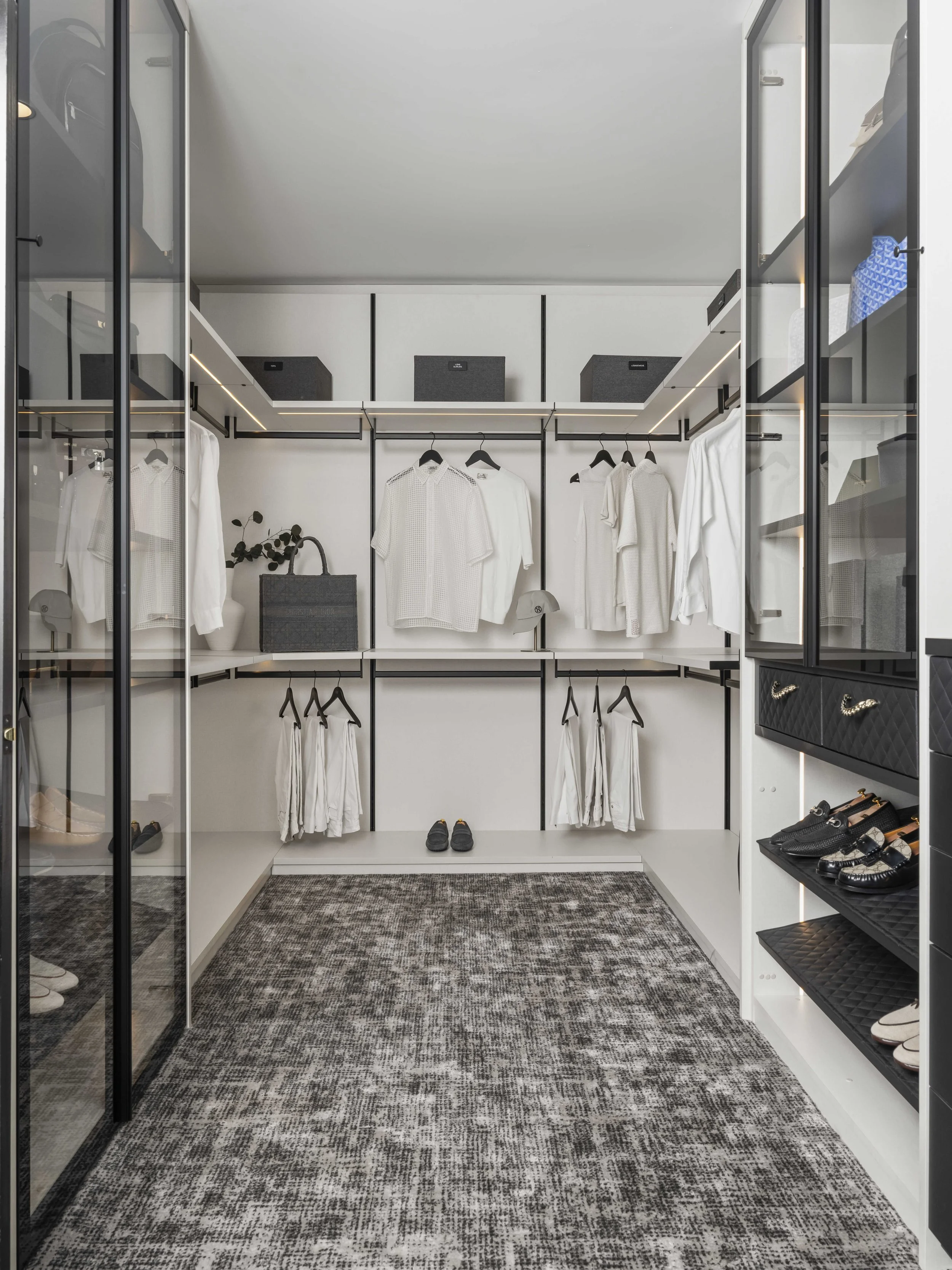A minimalist walk-in closet with white cabinetry, black hanging rods, and integrated LED shelf lighting. Features organized white shirts, a grey Dior tote, and a textured grey patterned carpet.
