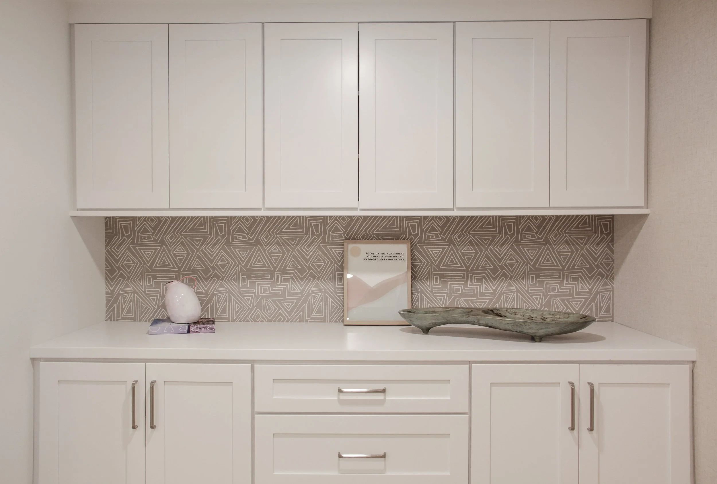 Built-in storage unit with white shaker-style cabinetry featuring upper and lower cabinets with brushed nickel hardware, white quartz countertop, and geometric patterned backsplash in taupe with angular linear design, styled with white vase, books, a