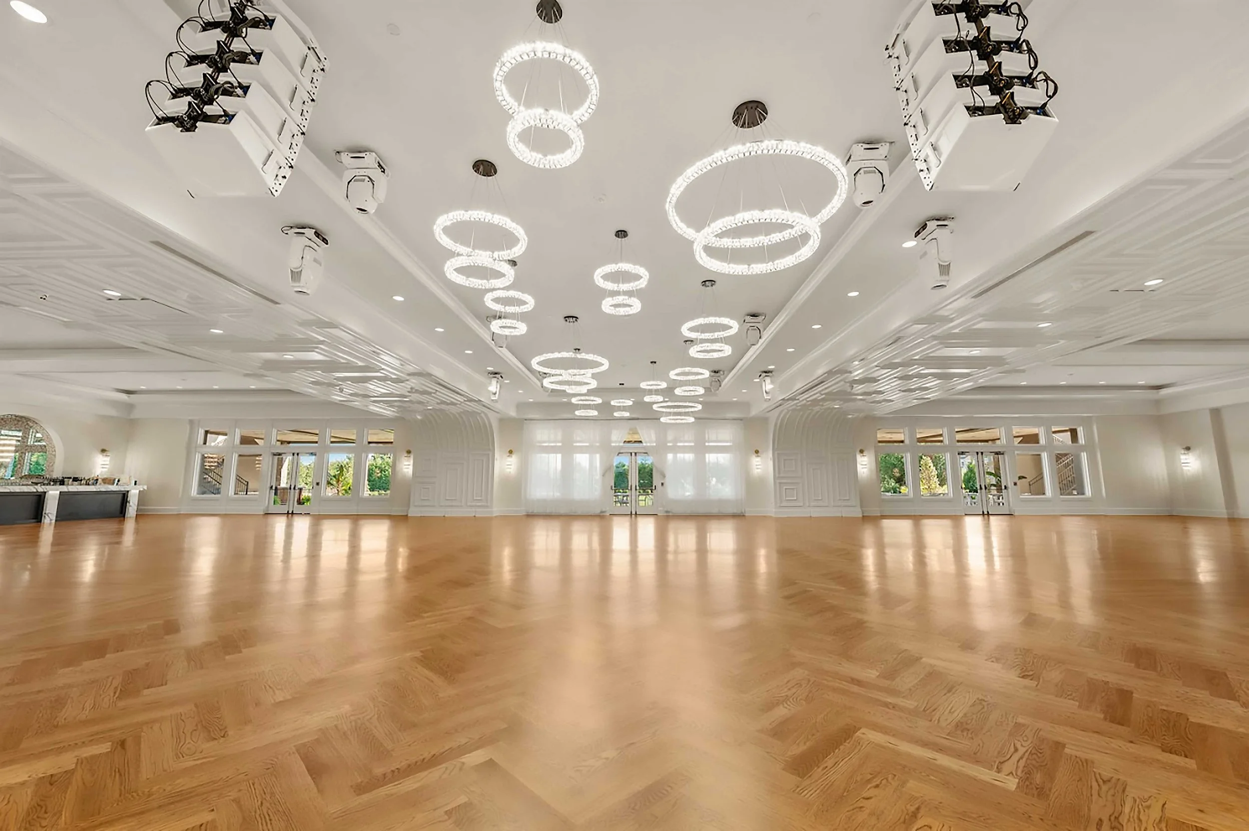 A wide-angle view of a massive, empty ballroom featuring light oak herringbone floors. The ceiling is adorned with numerous modern, glowing circular ring chandeliers, and the far wall consists of large windows letting in natural light.