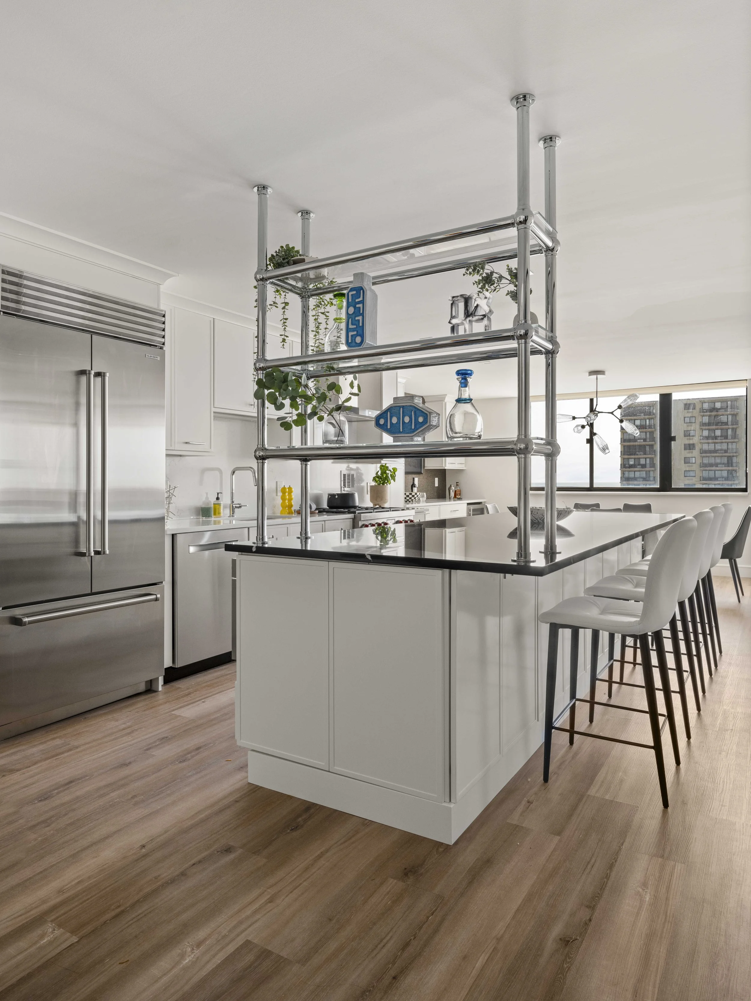 A kitchen island with a black countertop and white base, featuring a unique chrome industrial-style shelving unit used to display decorative blue glass and hanging plants.