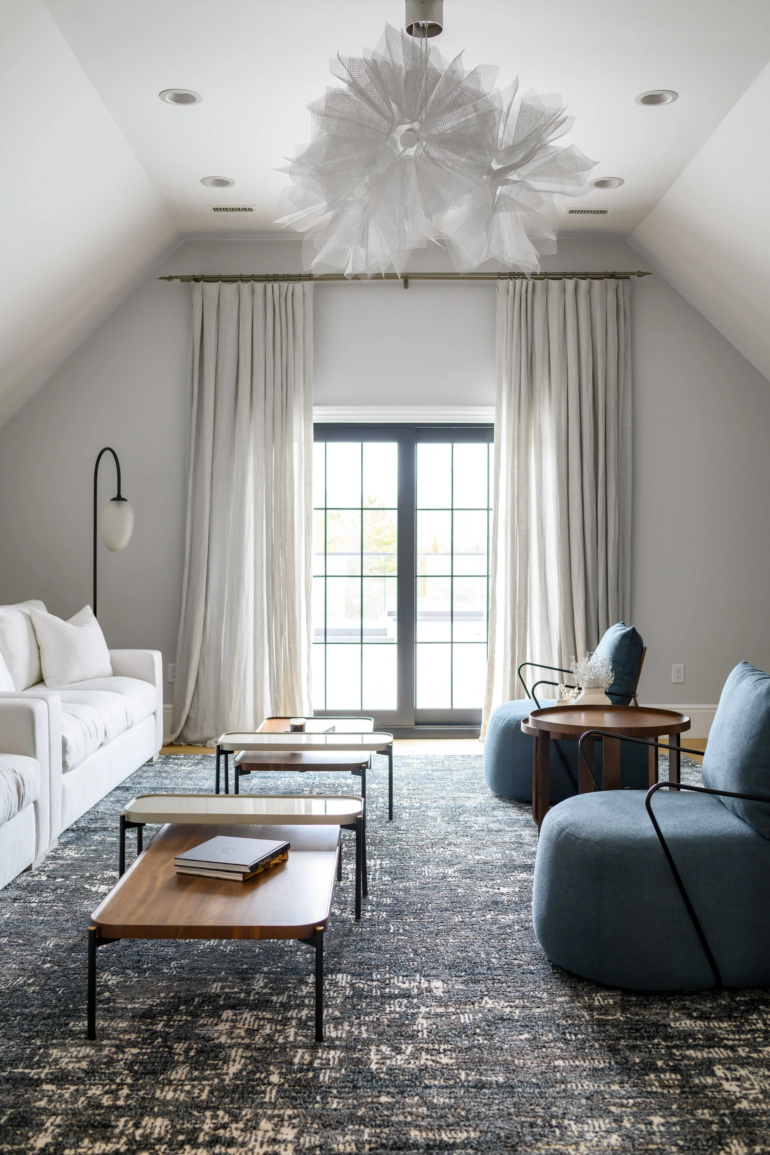 Vaulted living room with dramatic white tulle chandelier, blue upholstered chairs, nested wood and metal coffee tables, and black-framed French doors with natural light streaming through white curtains.