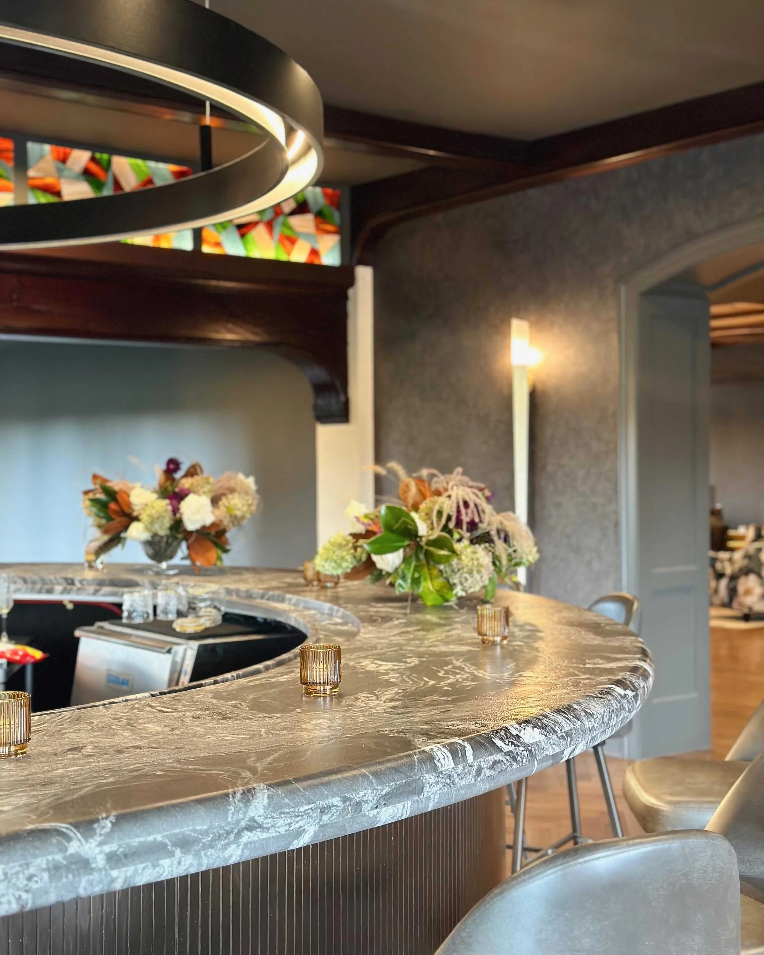 Close-up of a curved bar with a thick, grey veined marble top. In the background, a dark wooden architectural arch features a colorful stained-glass inlay above floral arrangements.