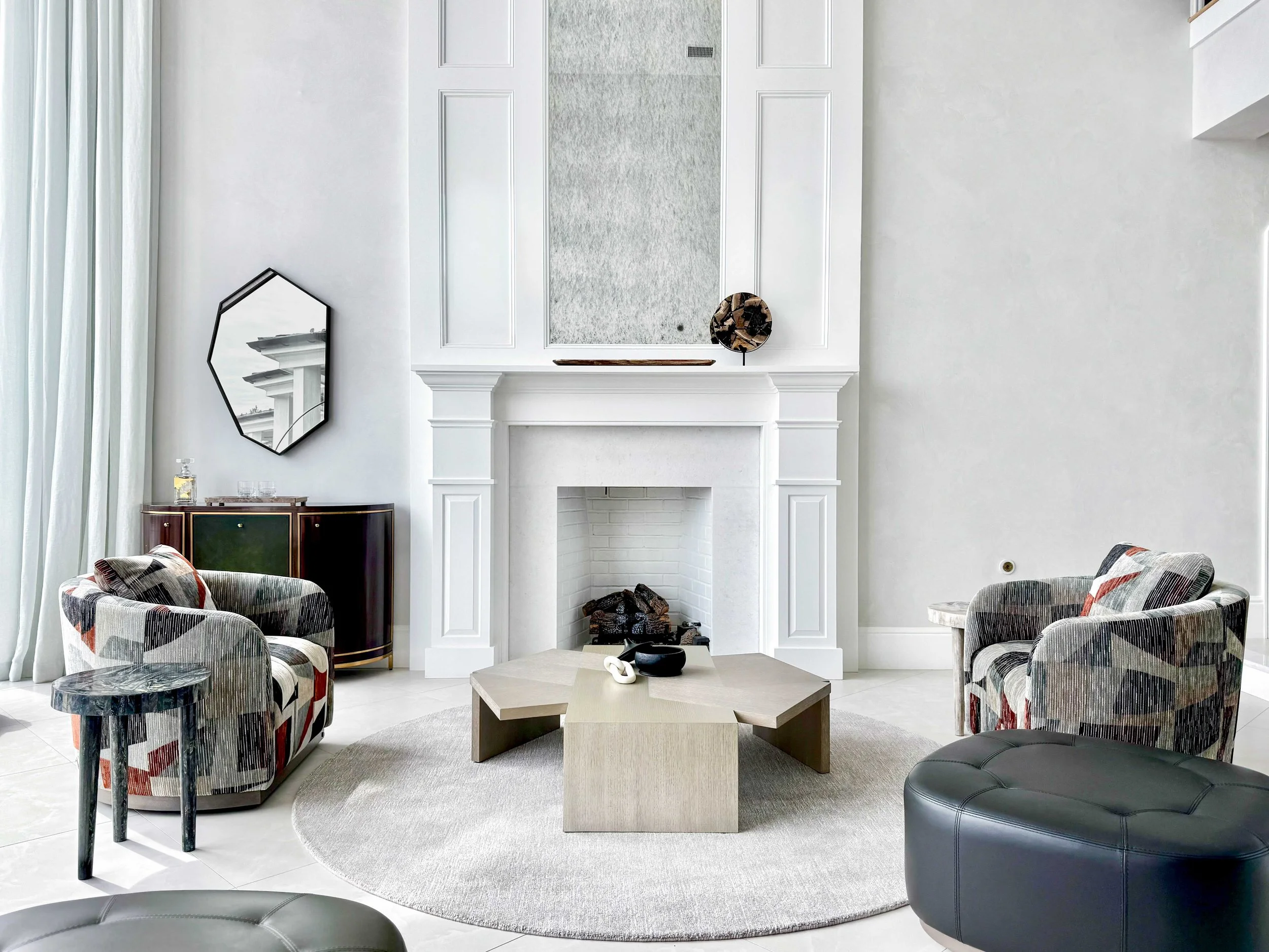 Bright living room with white traditional fireplace and painted brick surround, pair of modern swivel chairs in abstract patterned fabric, geometric cream coffee table with white chain link sculpture and black bowl, round area rug, and arched doorway