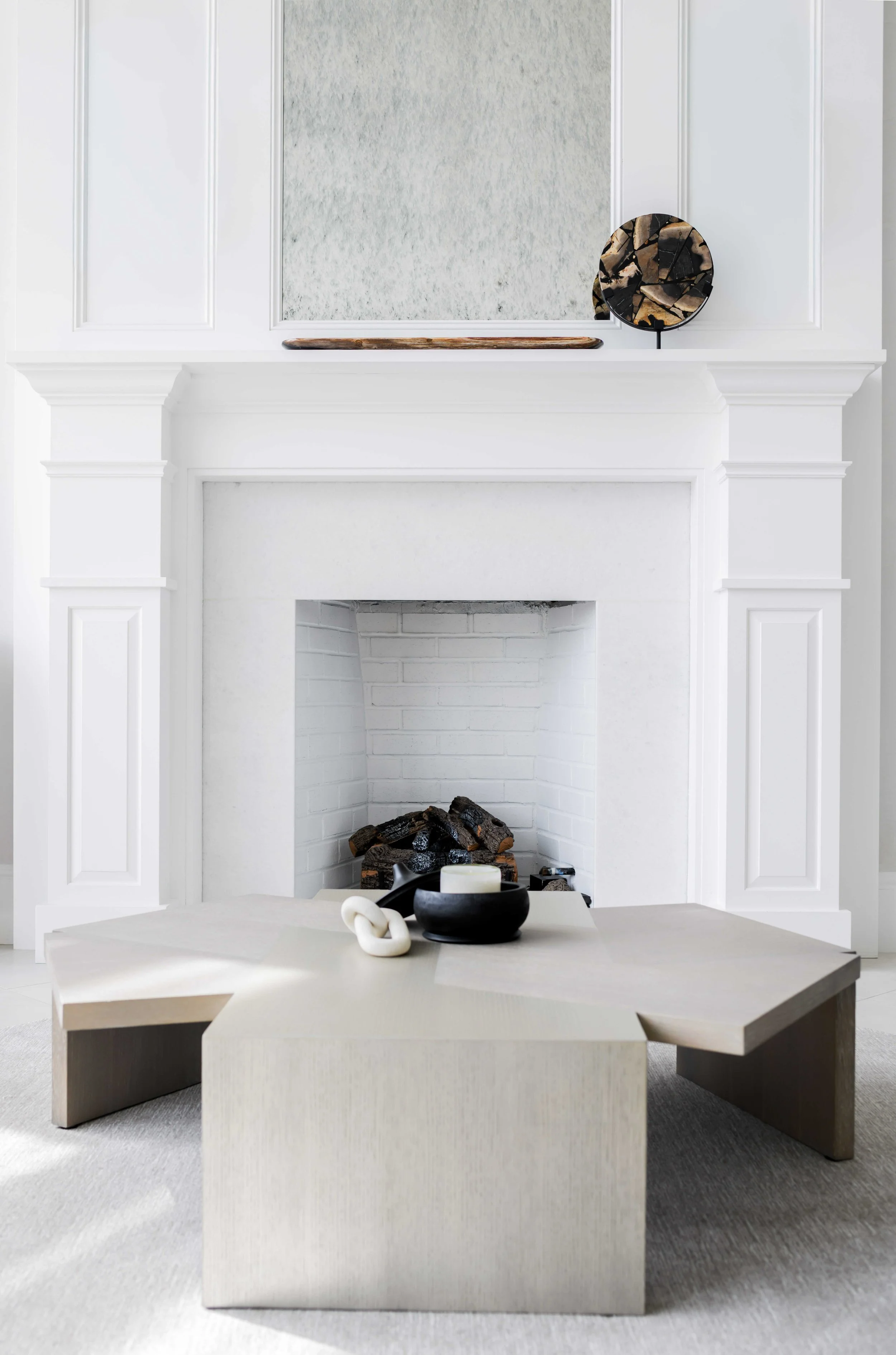 Minimalist fireplace vignette with white traditional surround and mantel, painted brick firebox with decorative logs, geometric cream coffee table with white chain link sculpture and black bowl, and live-edge wood tray with petrified wood disc on sta
