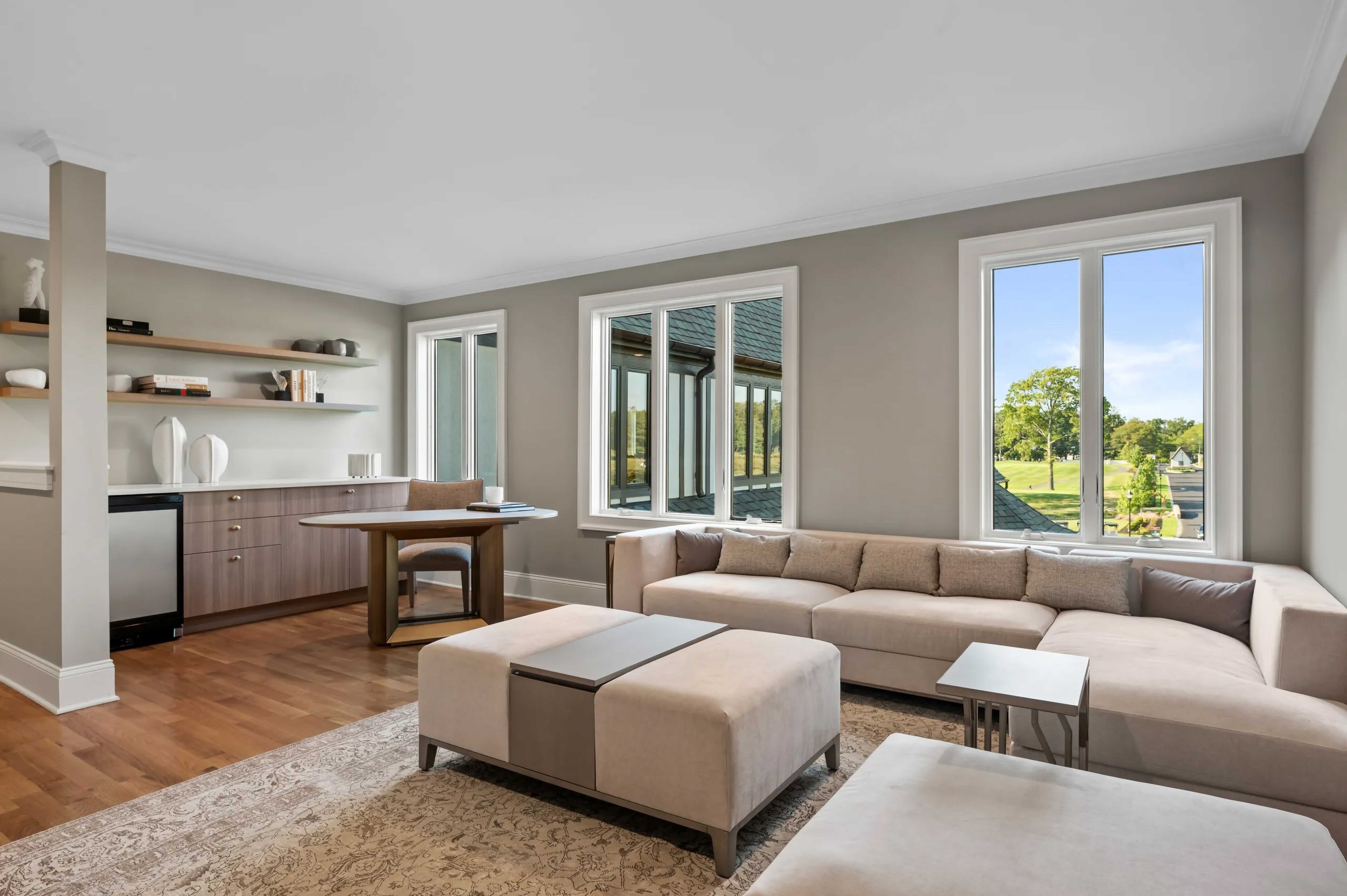 A bright, contemporary multi-use suite featuring a large beige L-shaped sectional sofa and matching oversized ottoman. In the background, there is a built-in wooden sideboard with white countertops, open shelving, and a small integrated refrigerator.