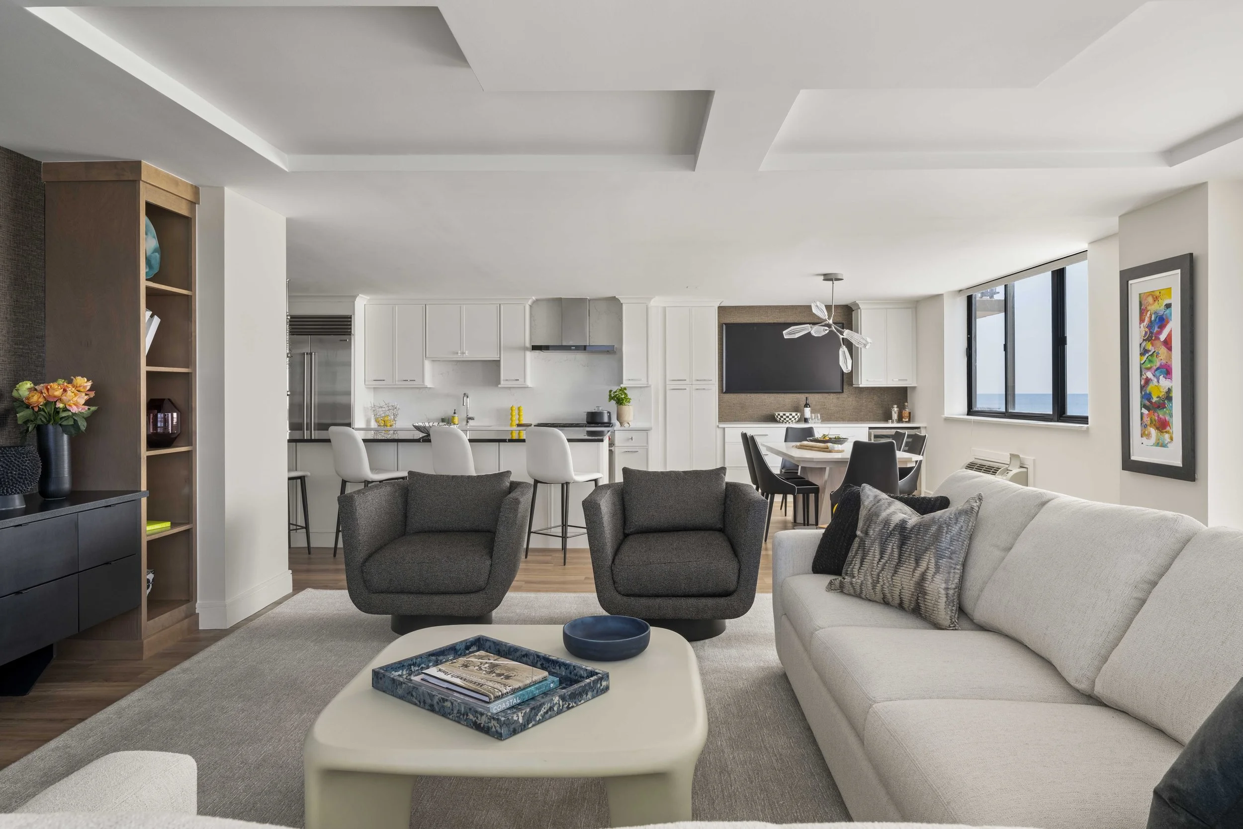 A wide view from the living room looking toward the kitchen and dining area. In the foreground are two charcoal gray armchairs and a cream-colored coffee table with a blue tray.