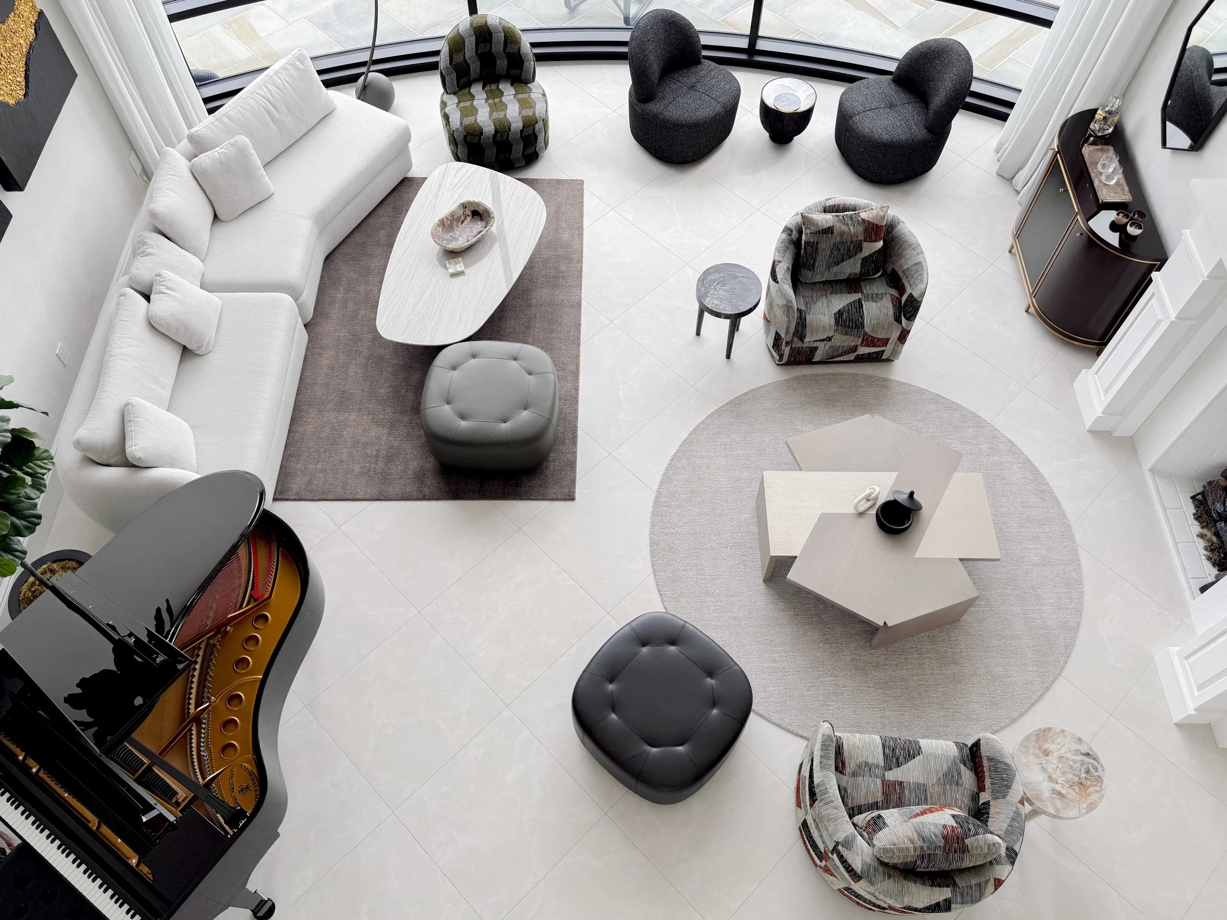 Aerial view of two-story living space with black grand piano, white sectional sofa, patterned swivel chairs in geometric fabric, gray tufted ottomans, sculptural white coffee table, round gray area rug, and large windows with lake views.