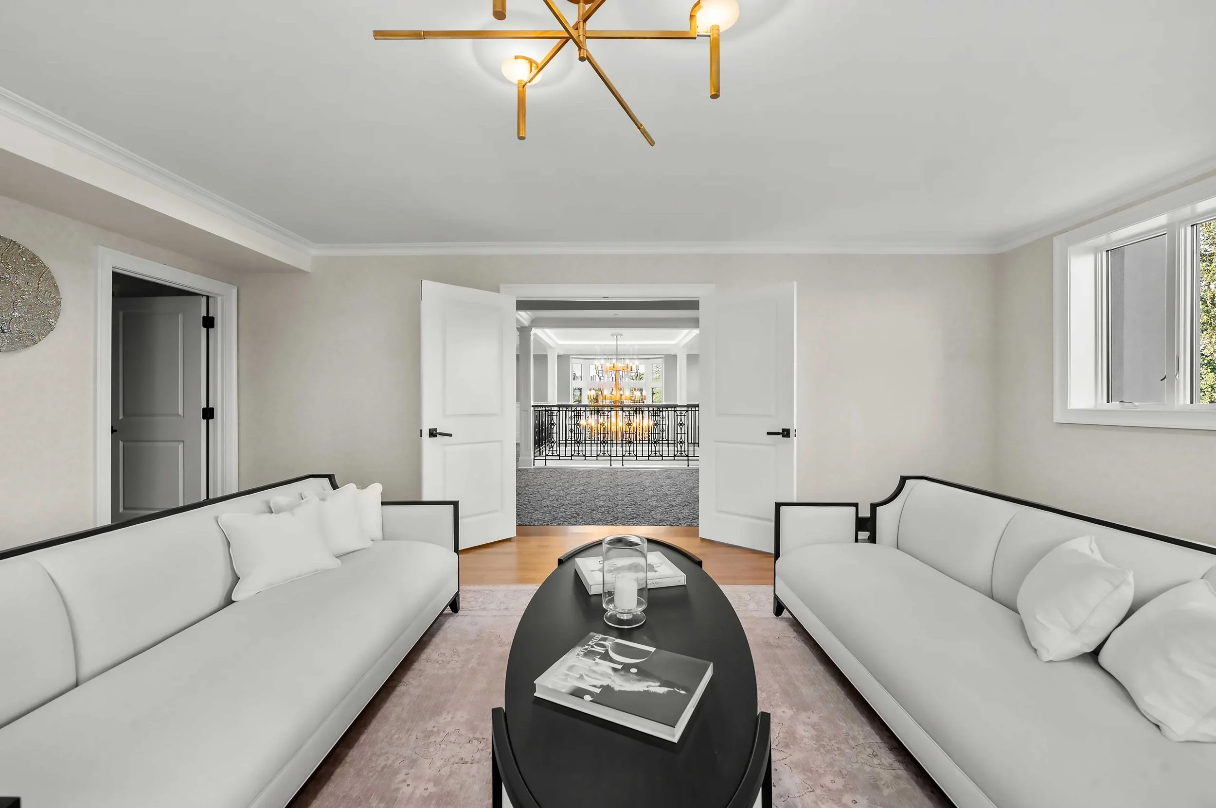 A bright, elegant lounge area with two long white sofas facing a black oval coffee table. A gold multi-arm chandelier hangs from the white ceiling, and open double doors in the background reveal a view of a grand hallway with a large chandelier and a