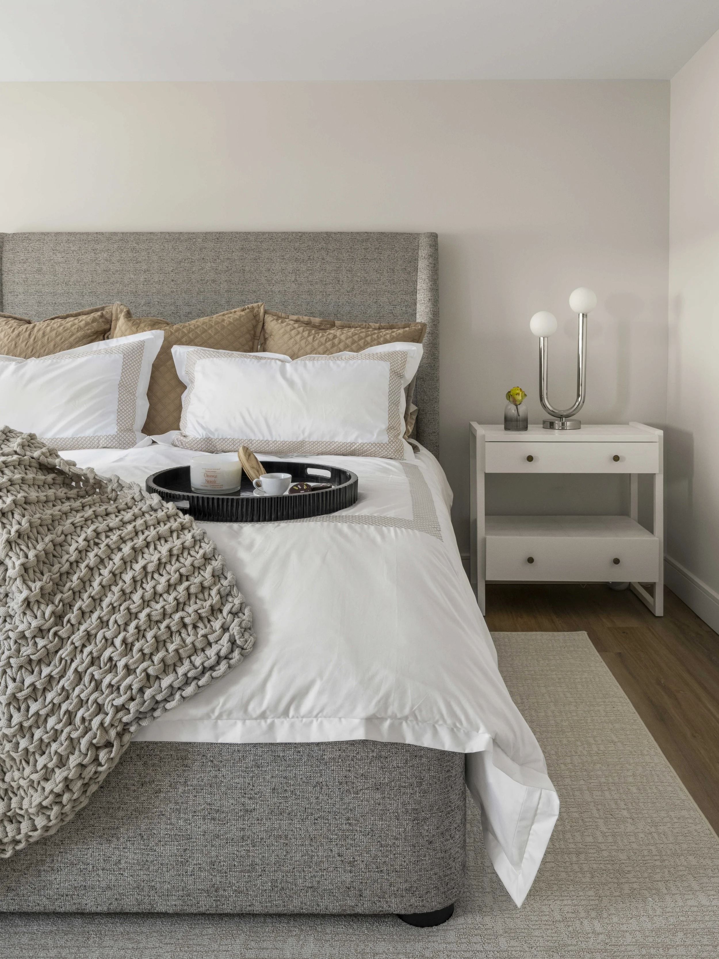 A close-up view of a bed with white and tan pillows and a black decorative tray. Next to it is a white nightstand holding a modern silver lamp with two globe bulbs and a small vase of flowers.
