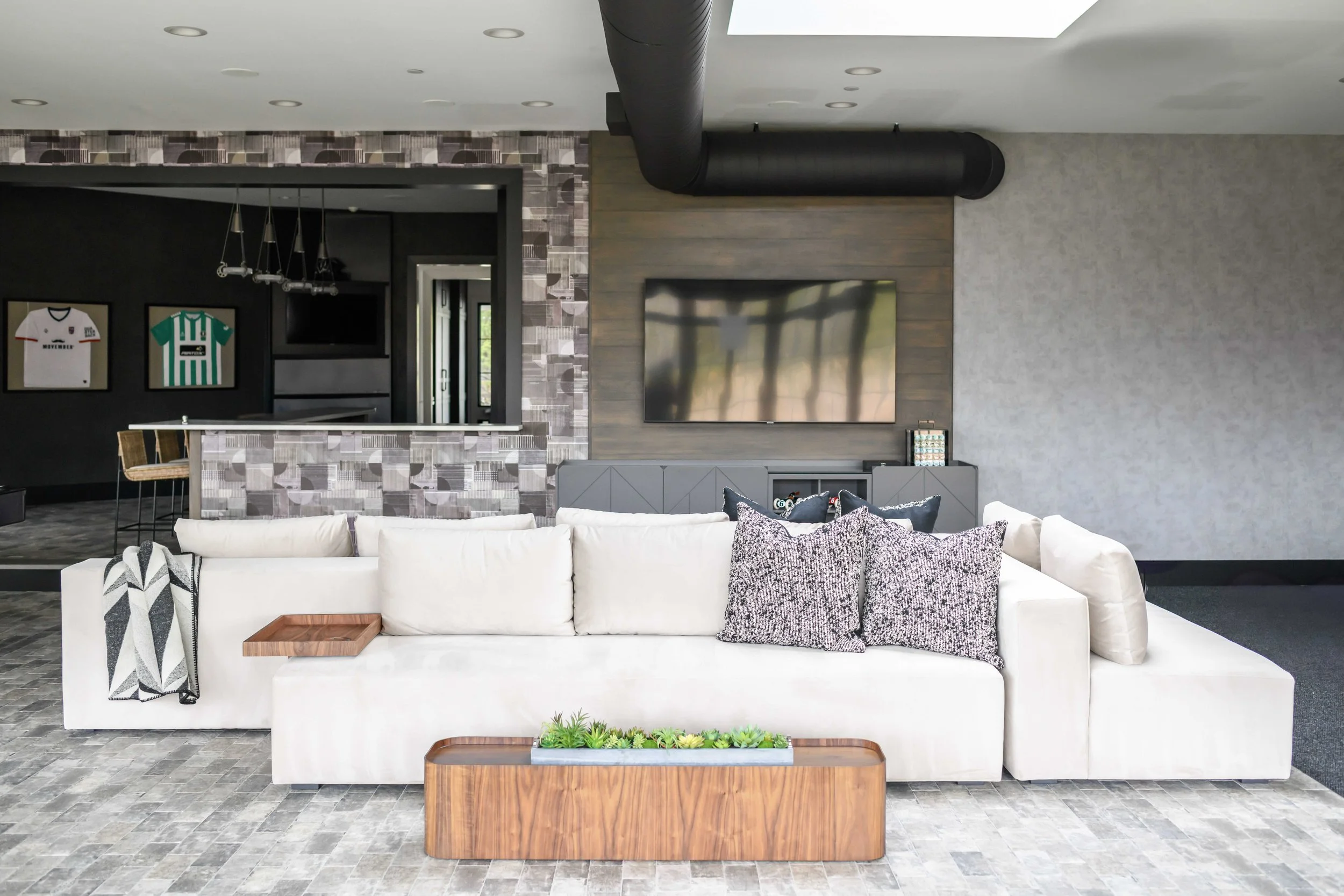 Modern entertaining space with white modular sectional, patterned throw pillows, wood coffee table with live-edge bowl and succulent planter, gray geometric tile fireplace surround, and bar area with framed sports jerseys.