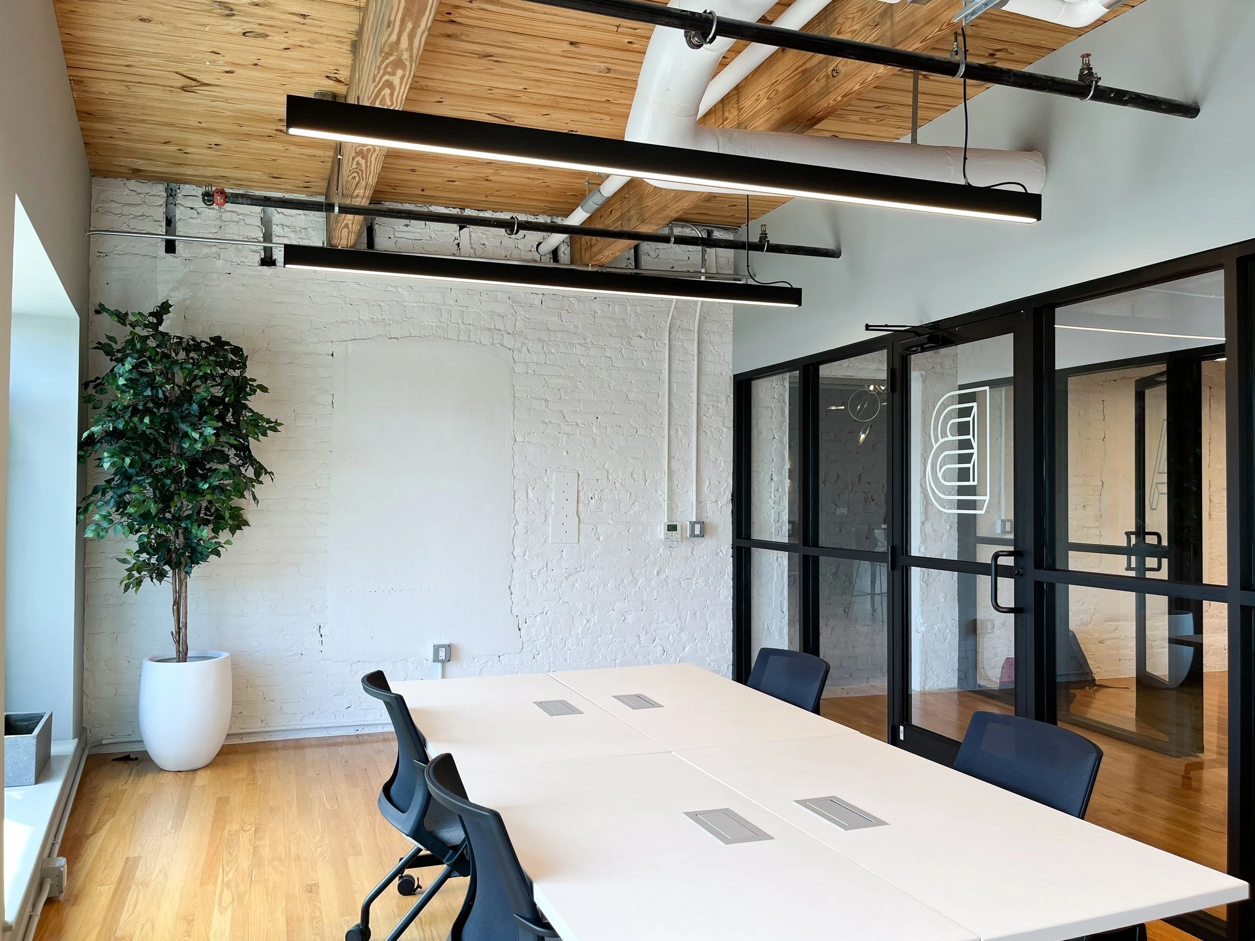 Meeting space with vaulted wood ceiling, painted white brick wall, white conference table with black chairs, black-framed glass sliding doors with company logo, black linear pendant lighting, large potted ficus tree, wood flooring, and exposed indust