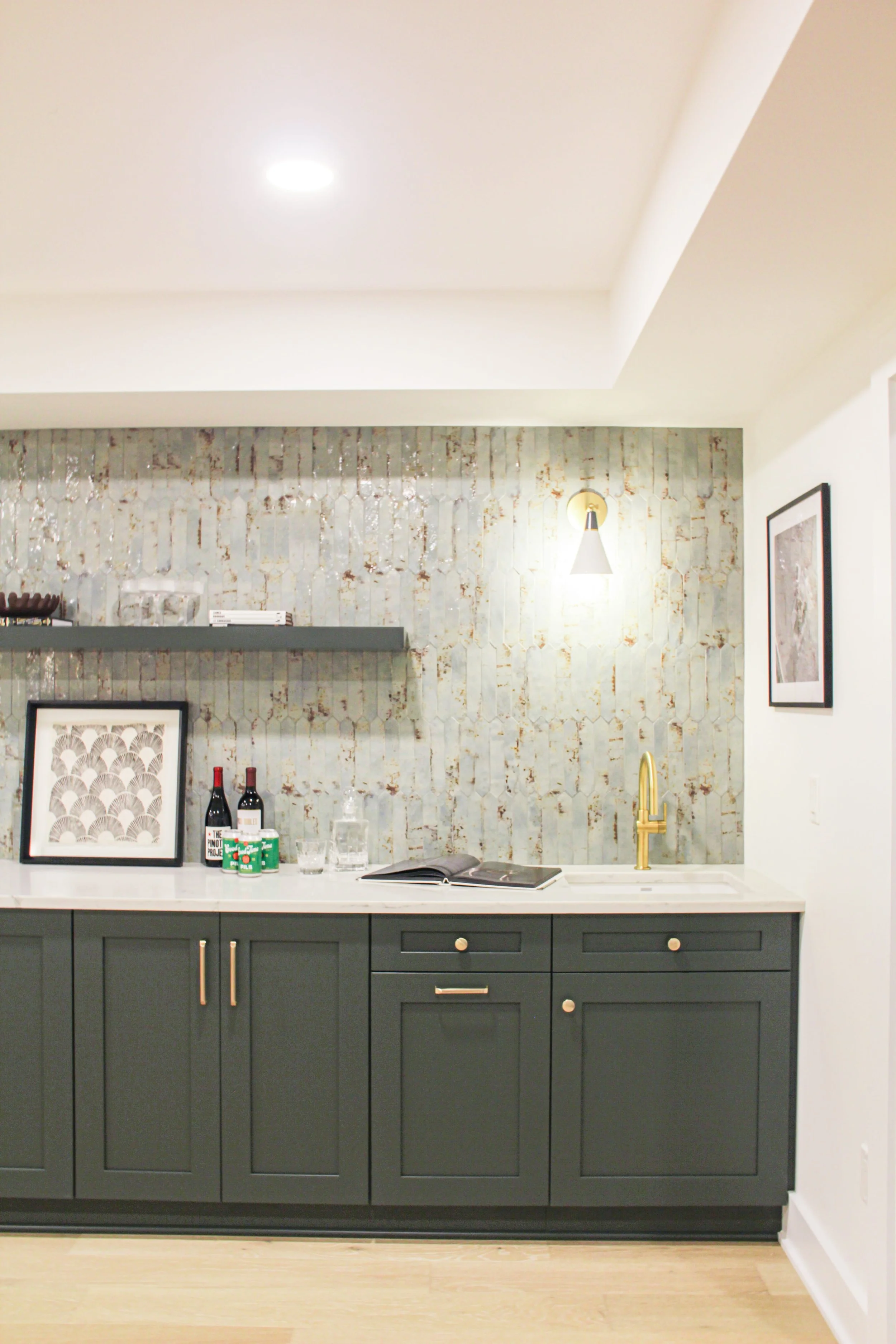 Coffered ceiling detail, shaker cabinets with brass knobs, white quartz countertop, distressed white patina tile backsplash, floating shelf with framed art and wine bottles, and brass sconce and faucet accents.