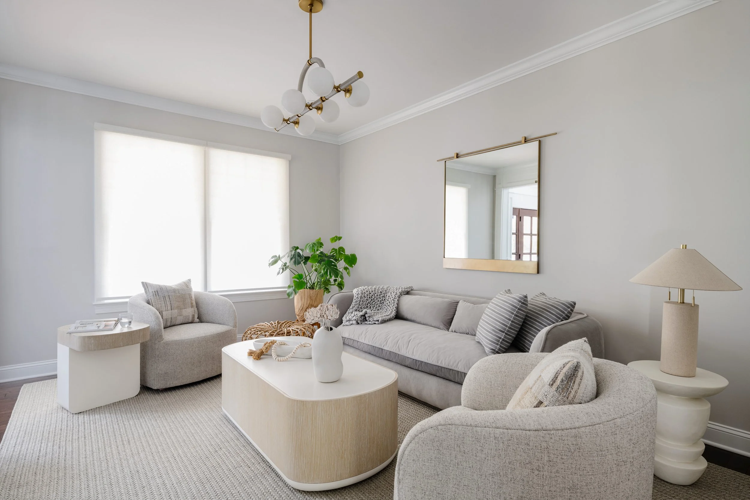 Serene living room in soft gray tones with contemporary brass and white glass globe chandelier, textured gray sofa with striped pillows, curved cream ottomans, wood-banded coffee tables, large brass-framed mirror, conical table lamp, potted plant, an