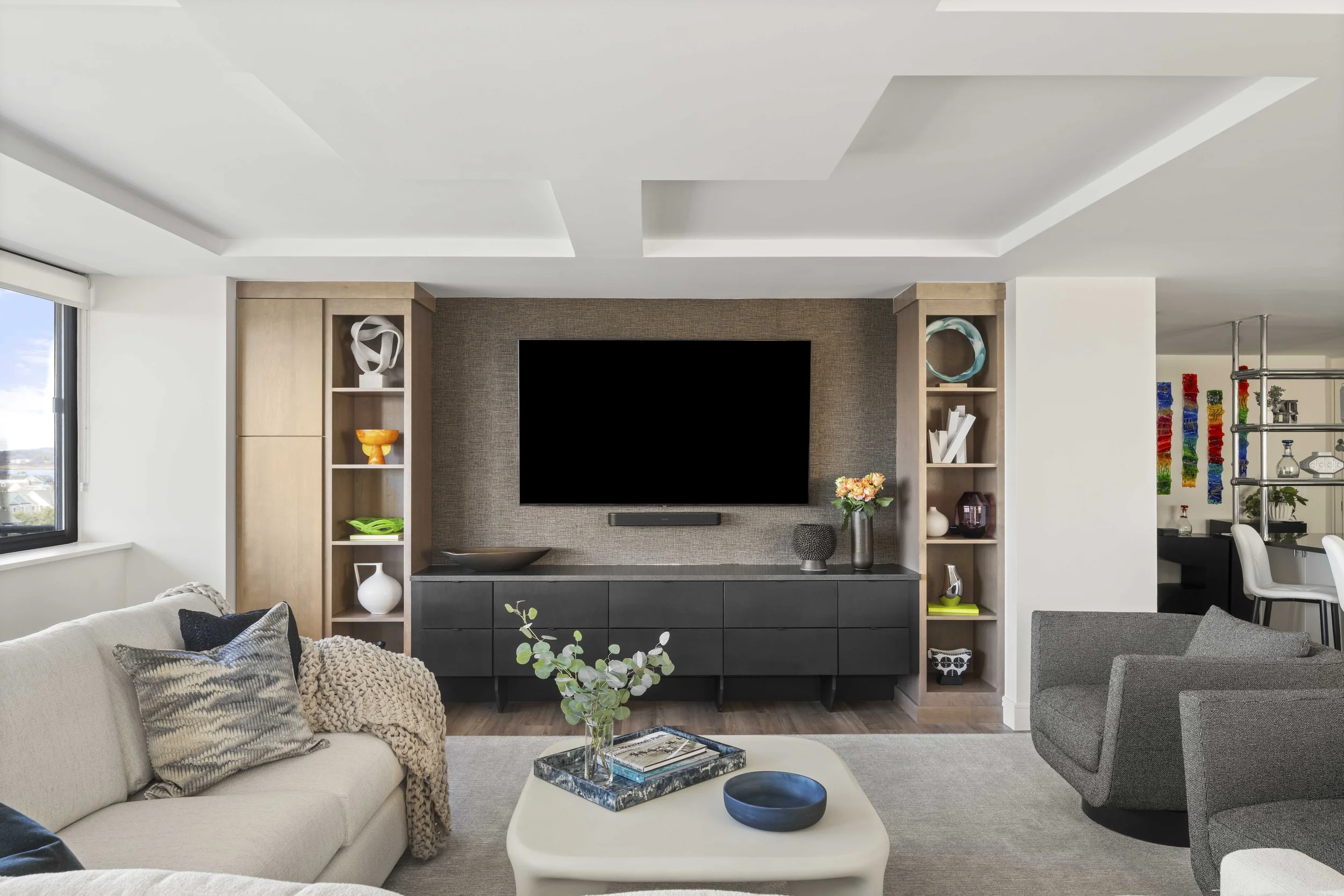 A modern entertainment center featuring a large wall-mounted TV on a brown textured accent wall. It is flanked by light wood built-in bookshelves filled with colorful art pieces and vases.