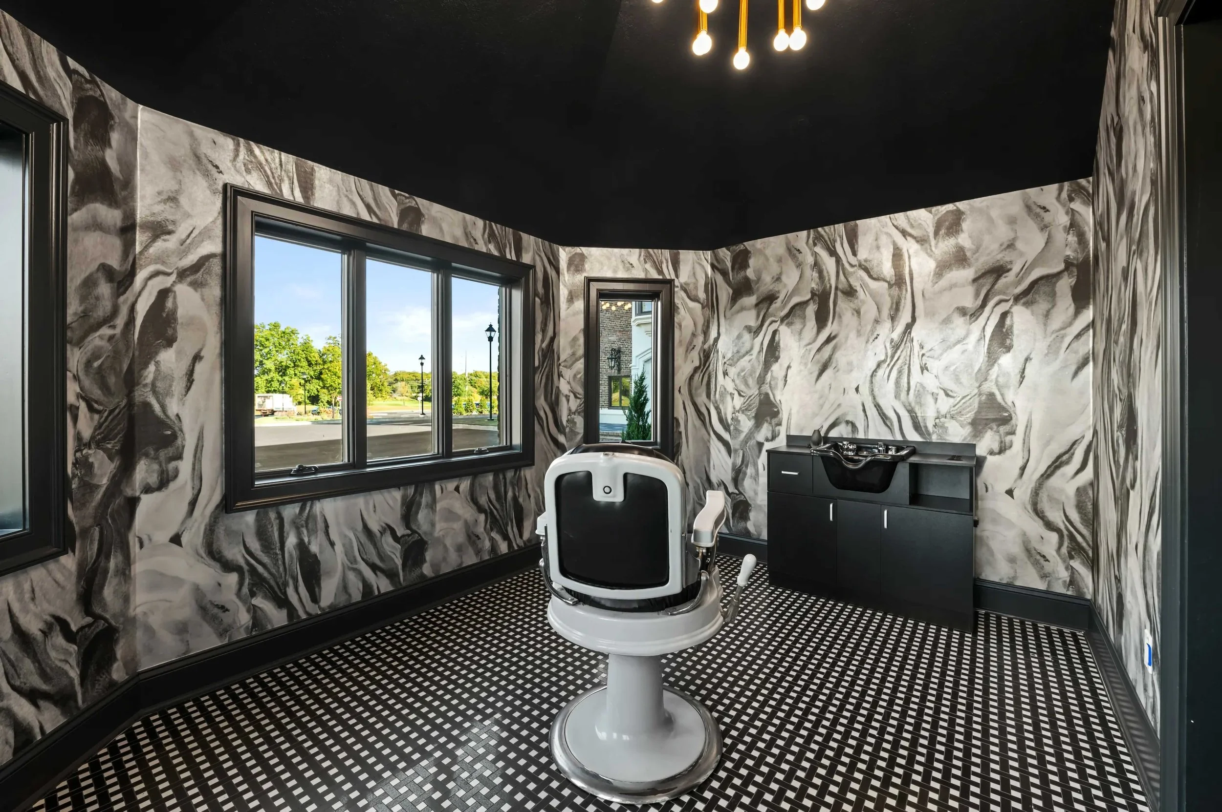 A boutique grooming room or barber suite featuring dramatic black-and-white marbled wallpaper and a black ceiling. A vintage-style white and black barber chair sits in the center of a black-and-white mosaic tile floor. The space includes a black cabi