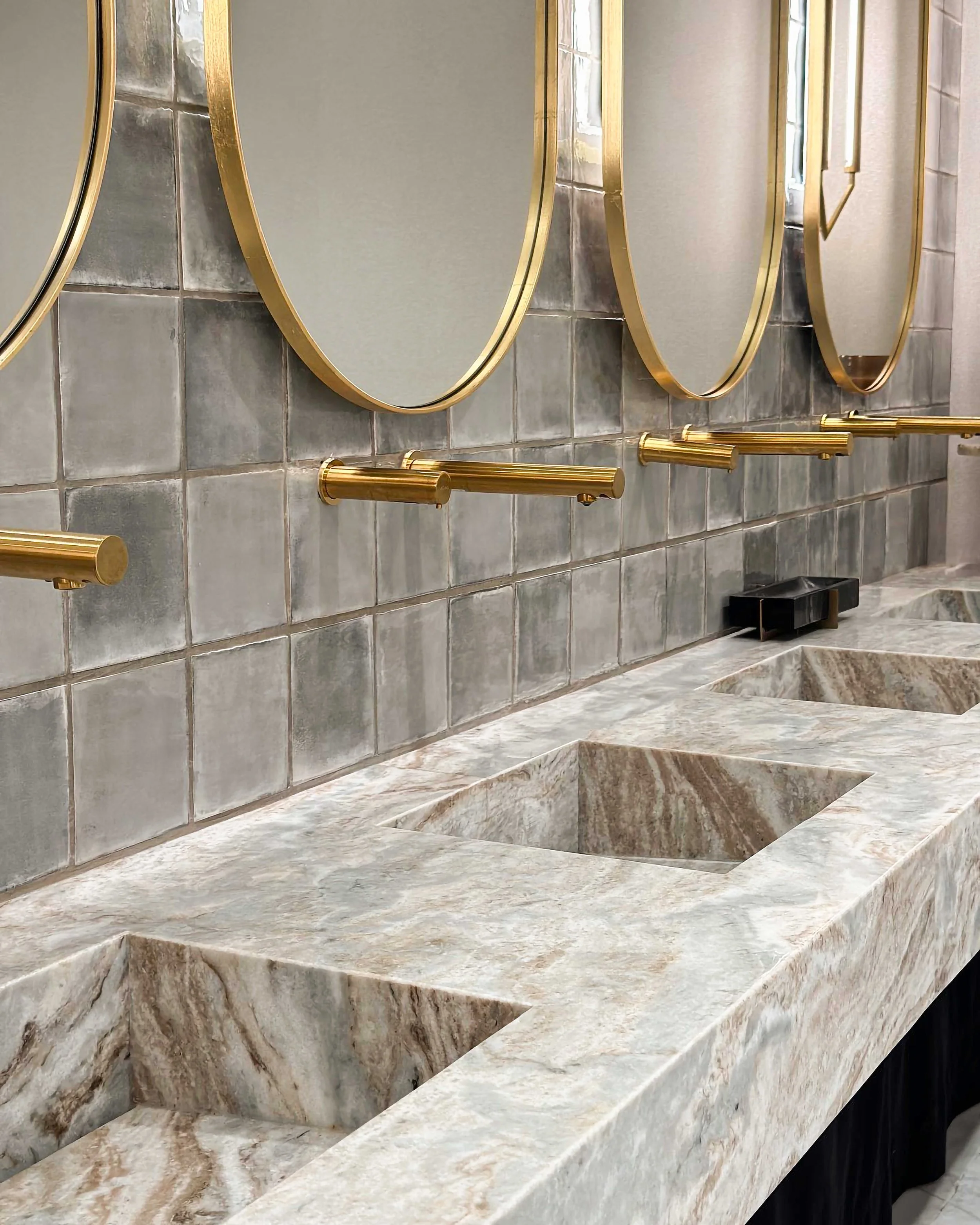 A luxury restroom featuring a long, continuous marble vanity with integrated square sinks. Gold-finished wall-mounted faucets match the large circular gold-framed mirrors set against grey square tiles.