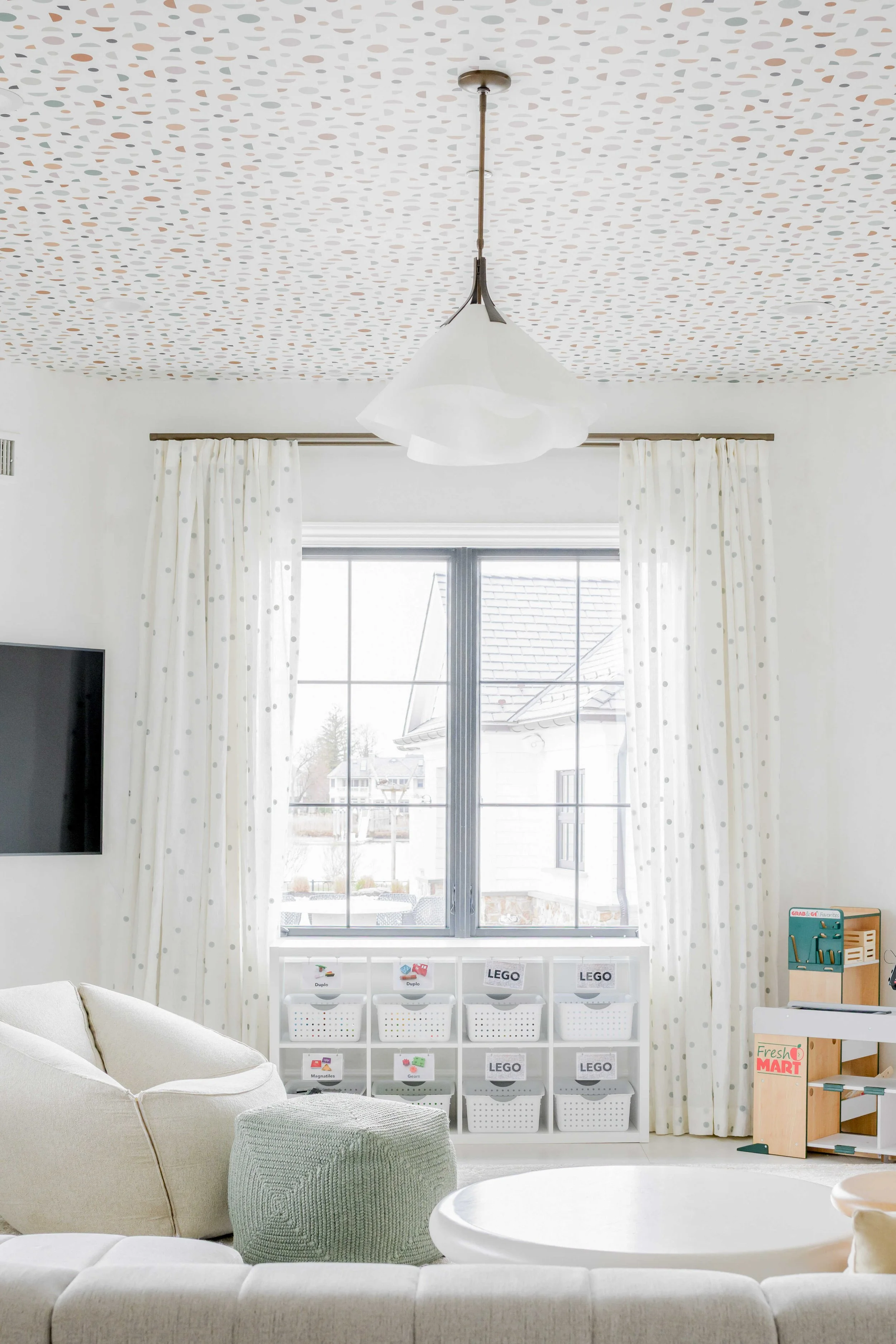 Bright children's playroom with playful ceiling wallpaper featuring scattered dots in peach, gray, and sage, white polka-dot curtains flanking window with toy storage cubes labeled for LEGO organization below, white pendant light, cream sectional sof