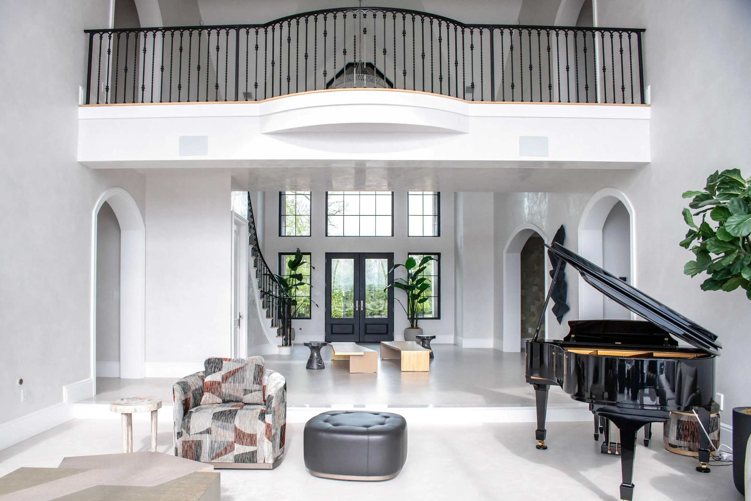 Grand two-story foyer with curved balcony featuring ornate black iron railing, black grand piano, arched doorways, black French entry doors with transom windows, modern sculptural seating, and oversized fiddle leaf fig plants.
