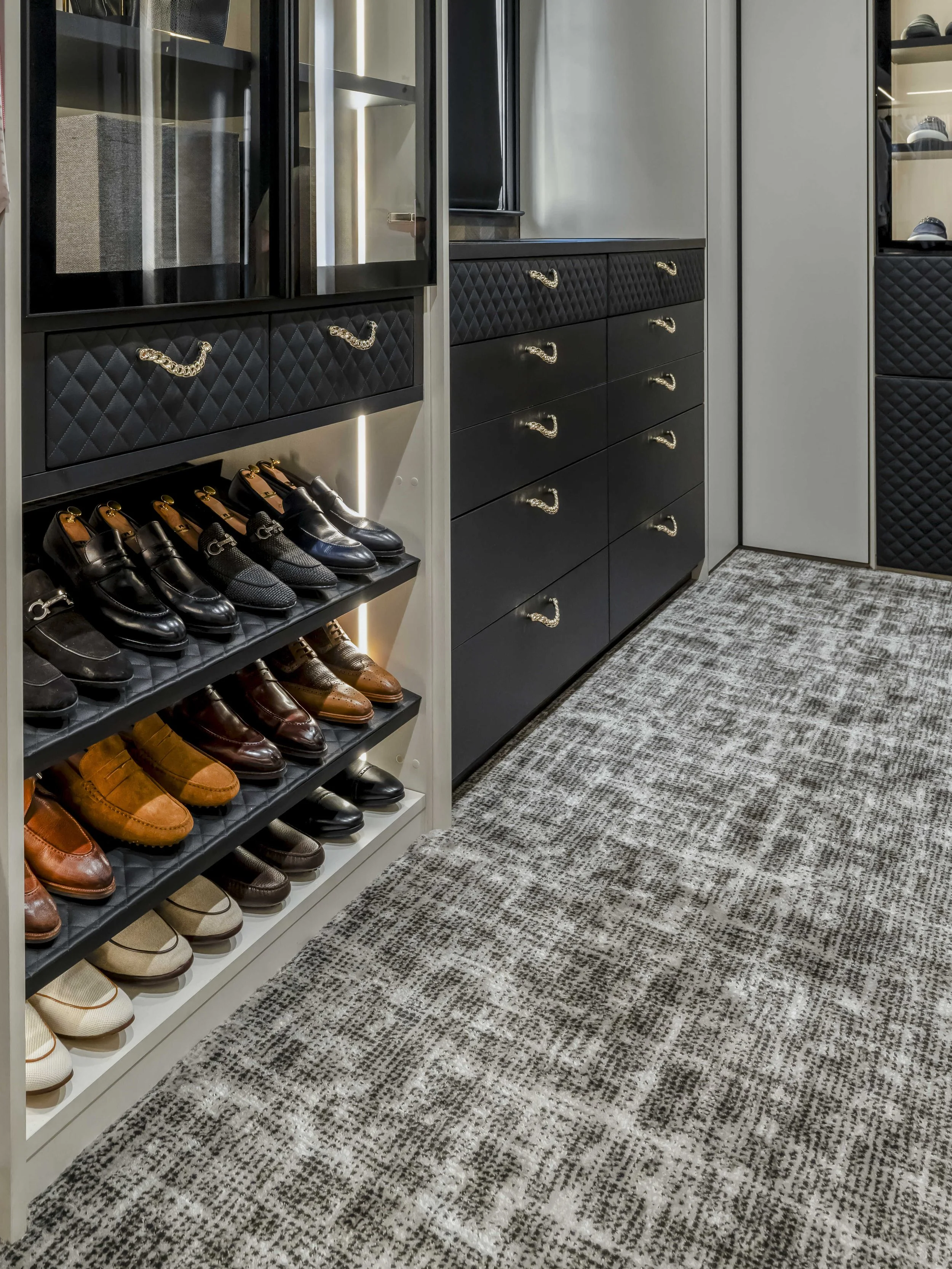 A low-angle shot of a luxury closet aisle featuring multiple rows of angled shoe shelves and black quilted drawers with gold hardware.