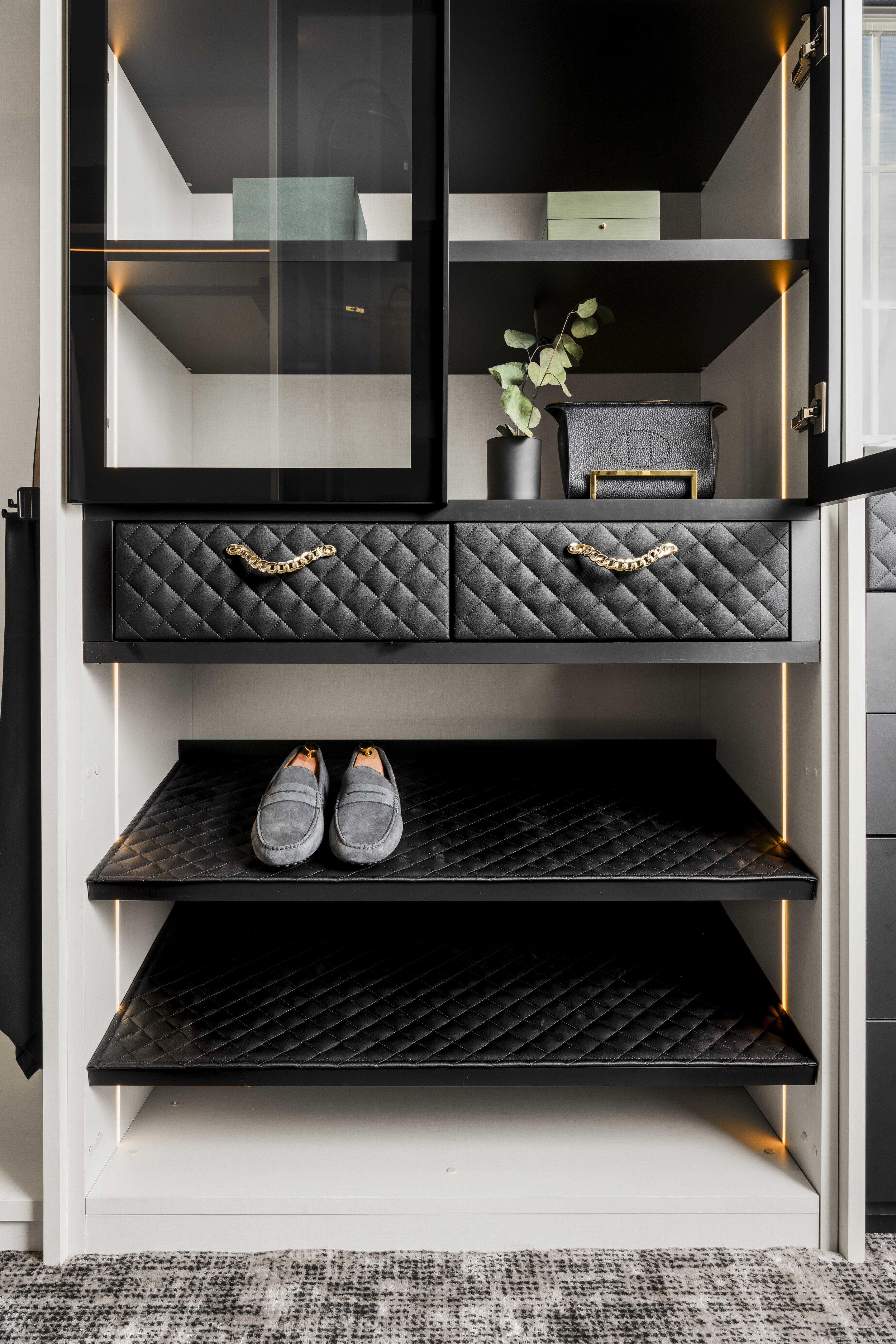 Close-up of custom closet shelving featuring black quilted leather drawer fronts with gold chain-link handles and LED-backlit shoe displays.