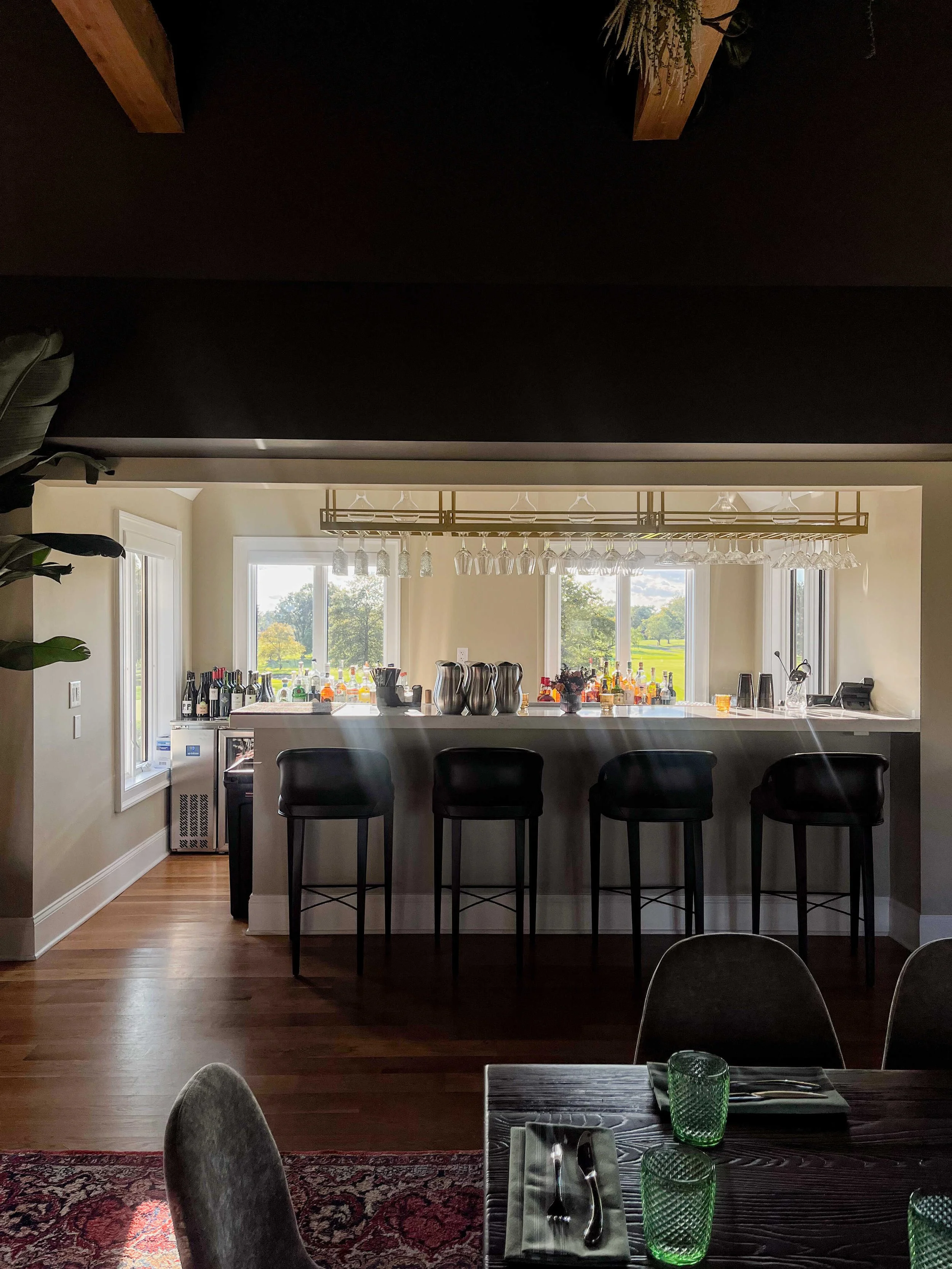 An indoor bar area with black leather barstools facing a white counter. Large windows behind the bar provide a bright view of a lush green golf course. A brass hanging glass rack and wooden ceiling beams add a rustic yet refined touch.