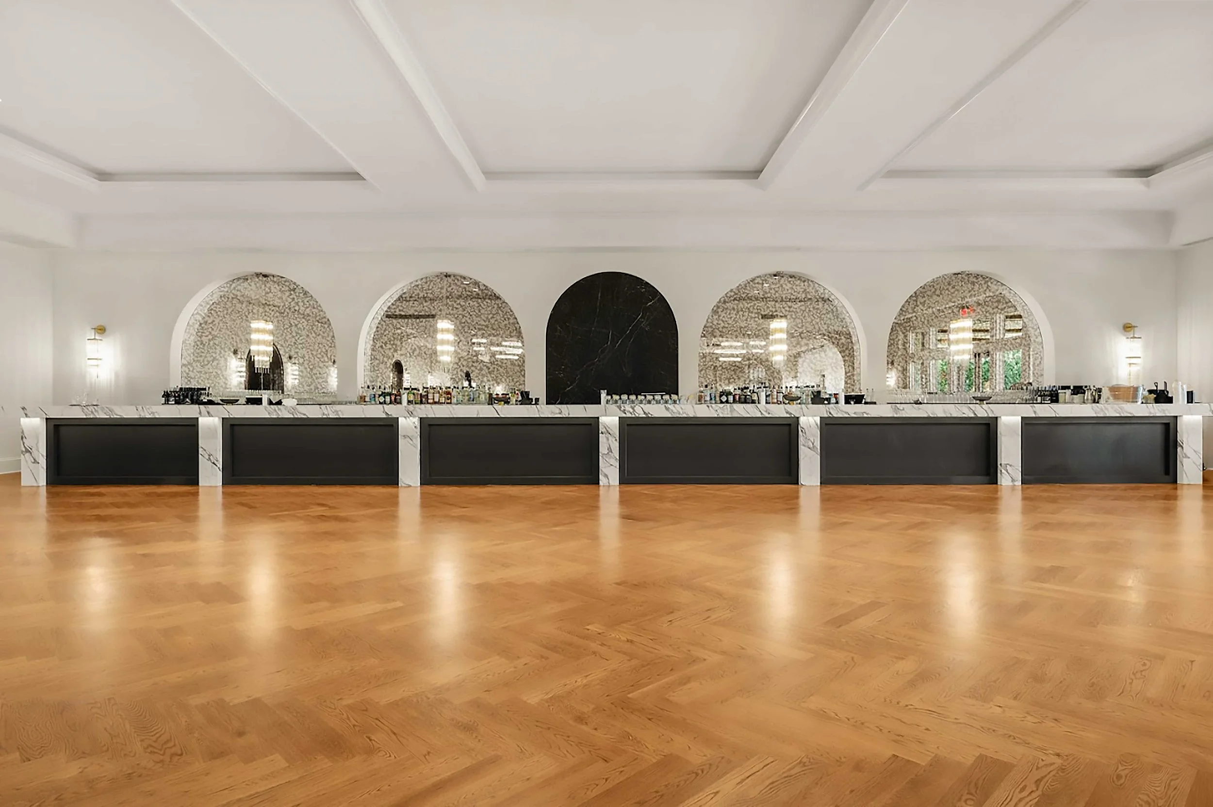 A long, modern bar area in the ballroom featuring a white marble countertop and dark paneled base. Behind the bar, five large arched mirrors reflect the room’s elegant lighting against a clean white wall.