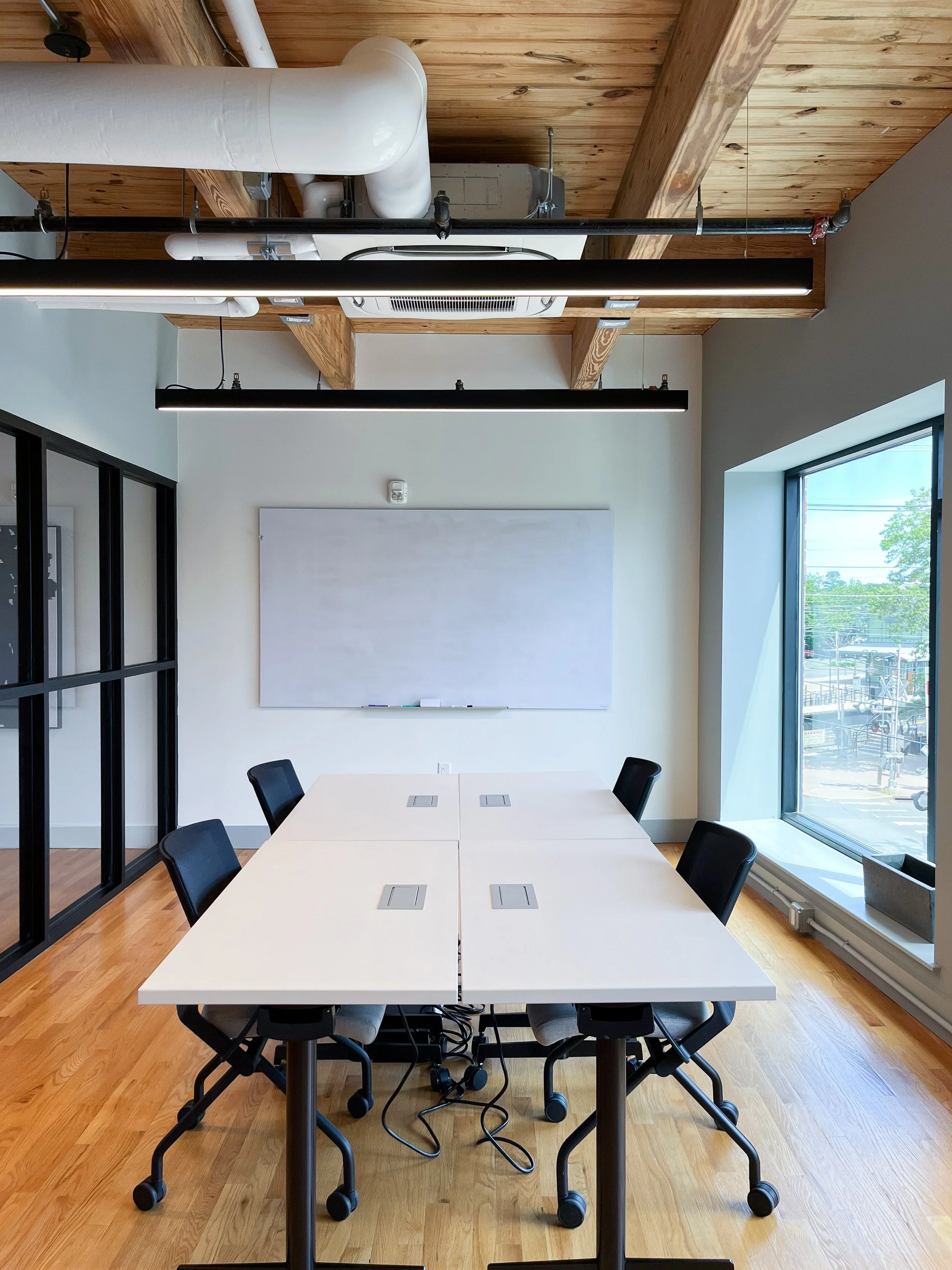 Conference room with natural wood ceiling and exposed beams, white walls with black-framed glass partitions, white modular meeting table with integrated power outlets, black rolling office chairs, wall-mounted whiteboard, large windows, and polished 