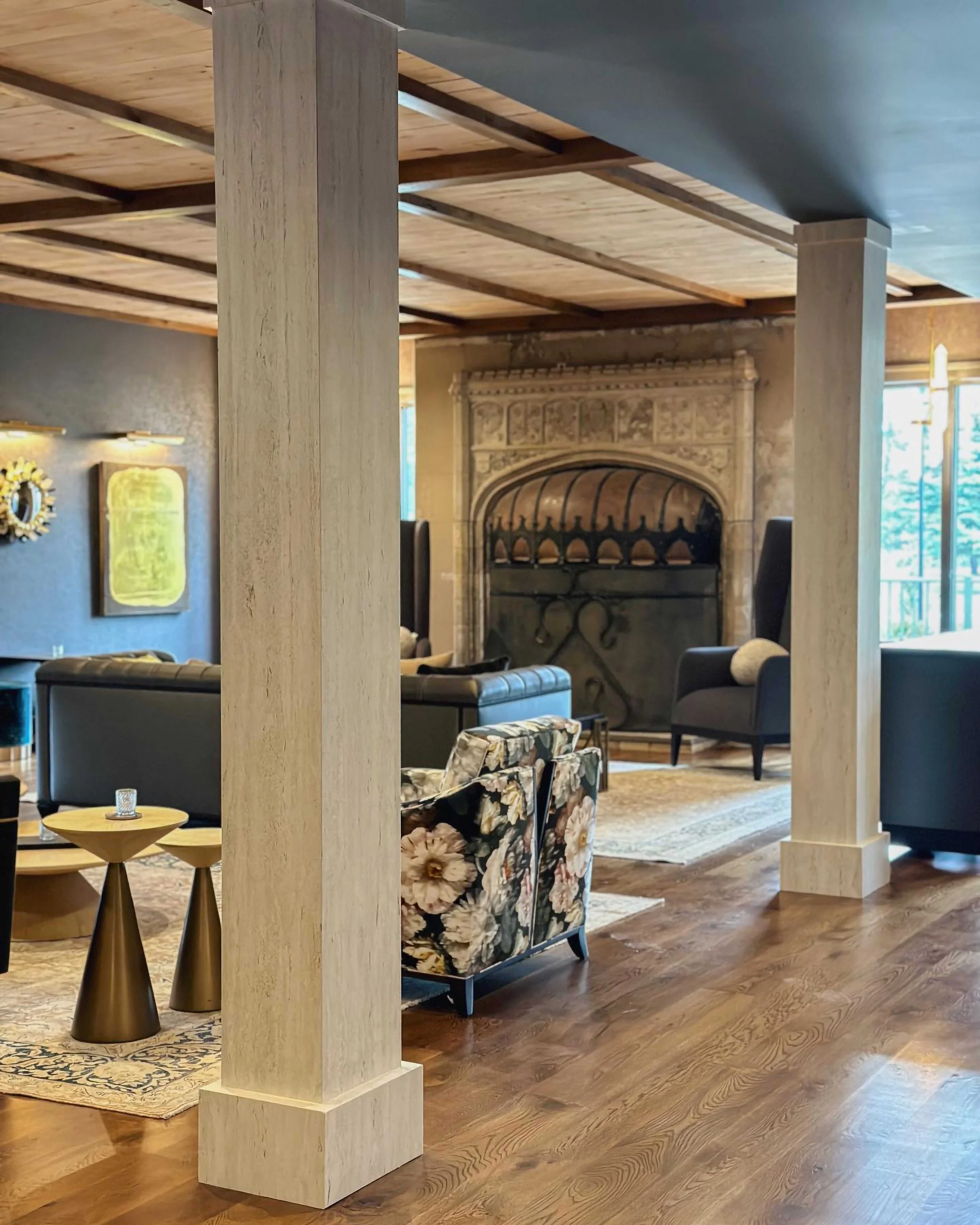 A cozy, upscale lounge featuring a large stone fireplace with intricate carvings. The room is furnished with dark leather sofas, floral-patterned armchairs, and mid-century modern side tables on a light-colored area rug.