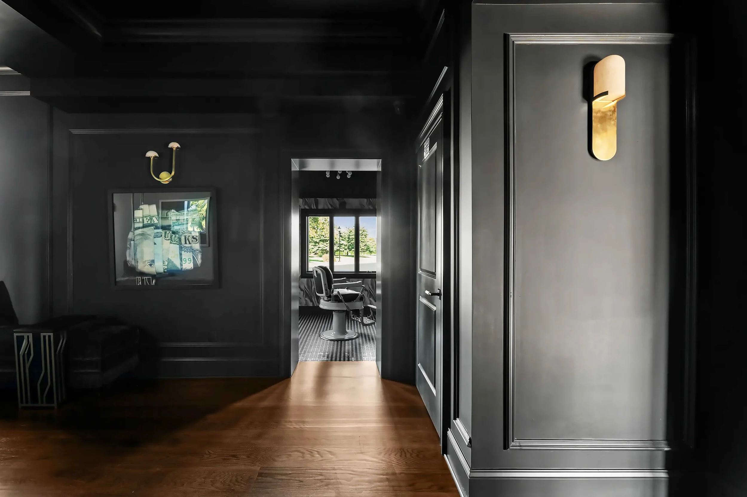 A moody, dark charcoal hallway featuring a modern gold wall sconce and a doorway leading into a bright barber suite.