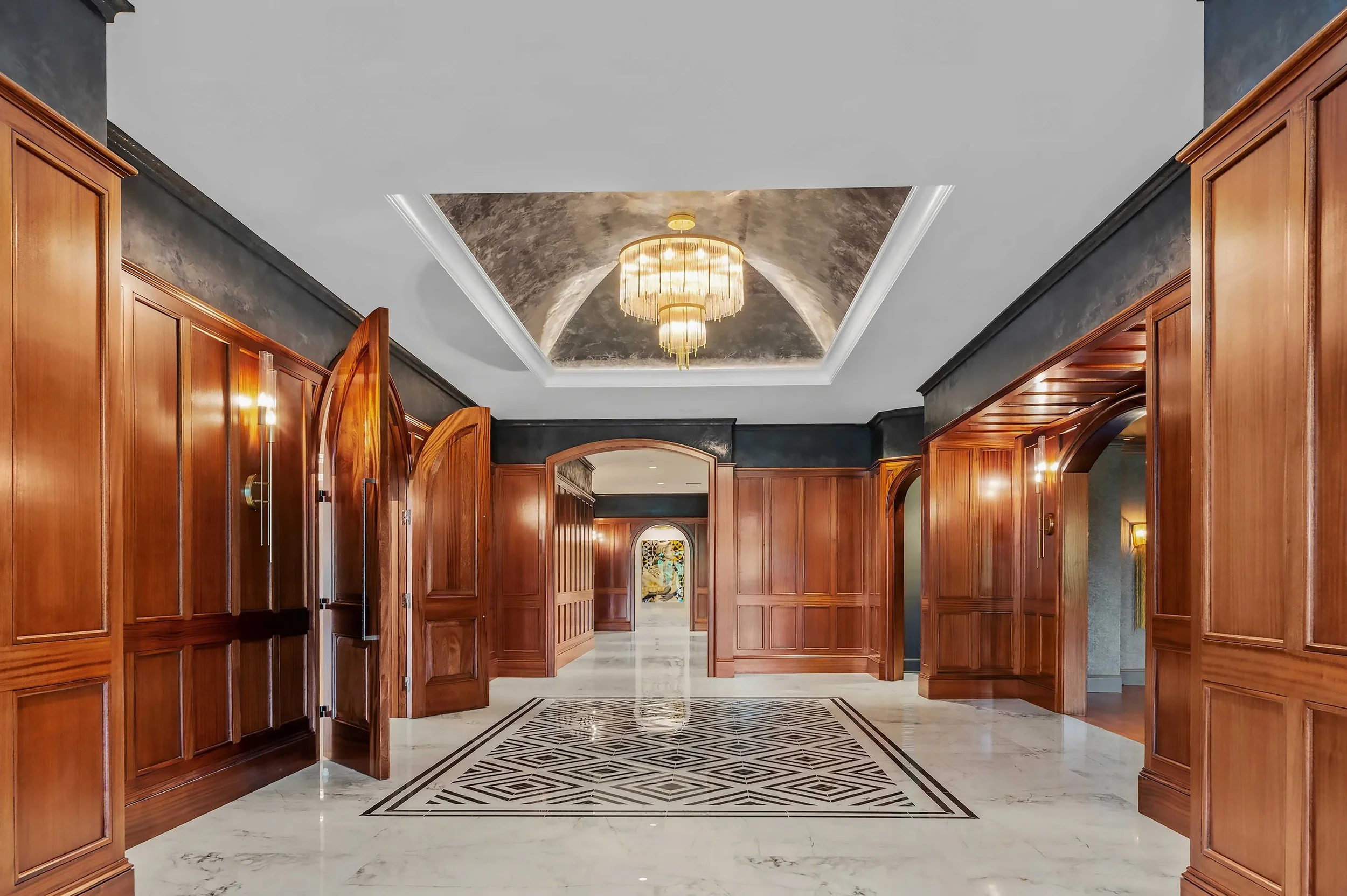 A grand entrance hall with rich wood paneling, arched doorways, a marble floor with geometric inlays, and a tiered crystal chandelier set in a domed ceiling.
