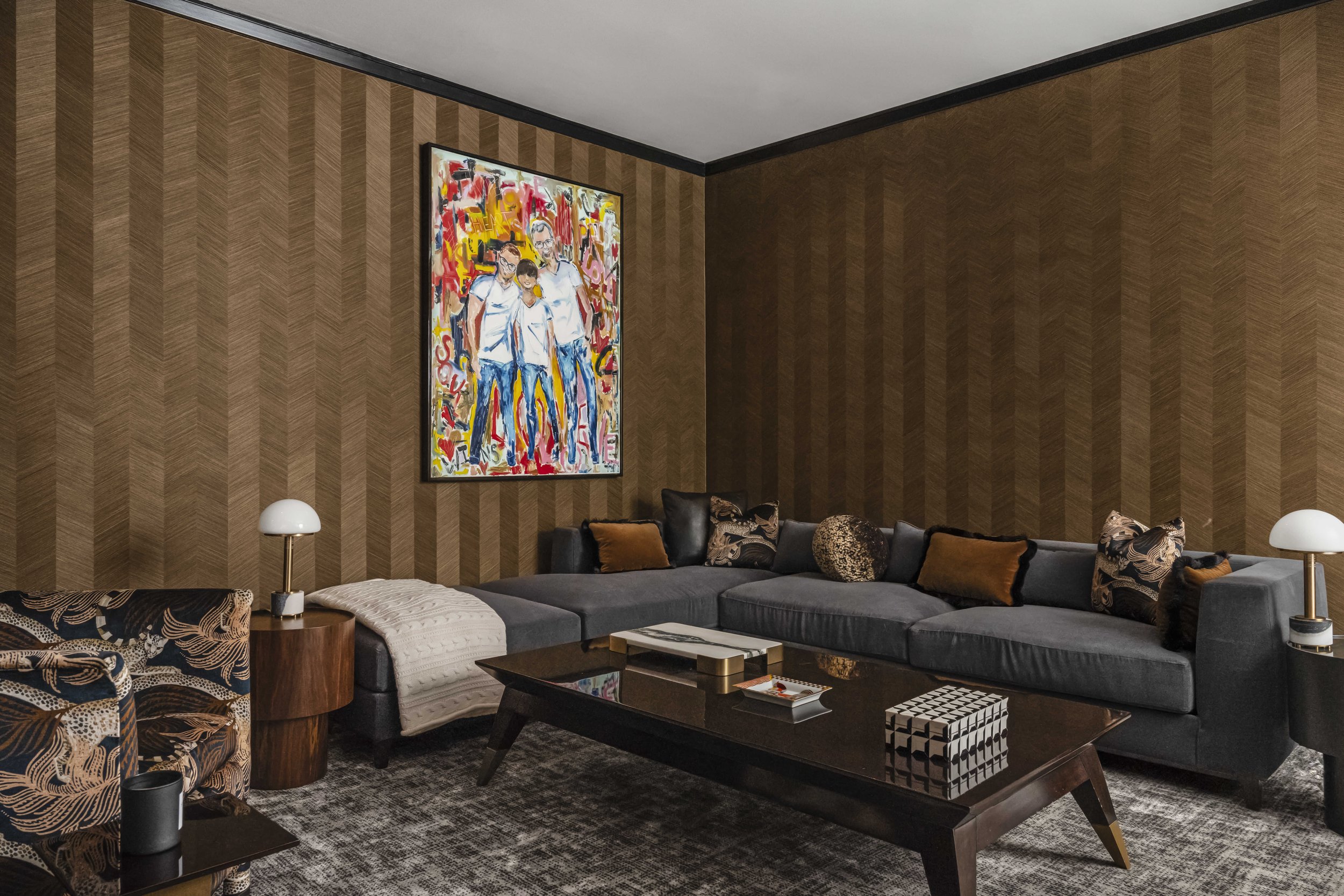 A sophisticated lounge area with gold-toned chevron wallpaper, a large grey sectional sofa, a dark wood coffee table, and a vibrant colorful art piece on the wall.