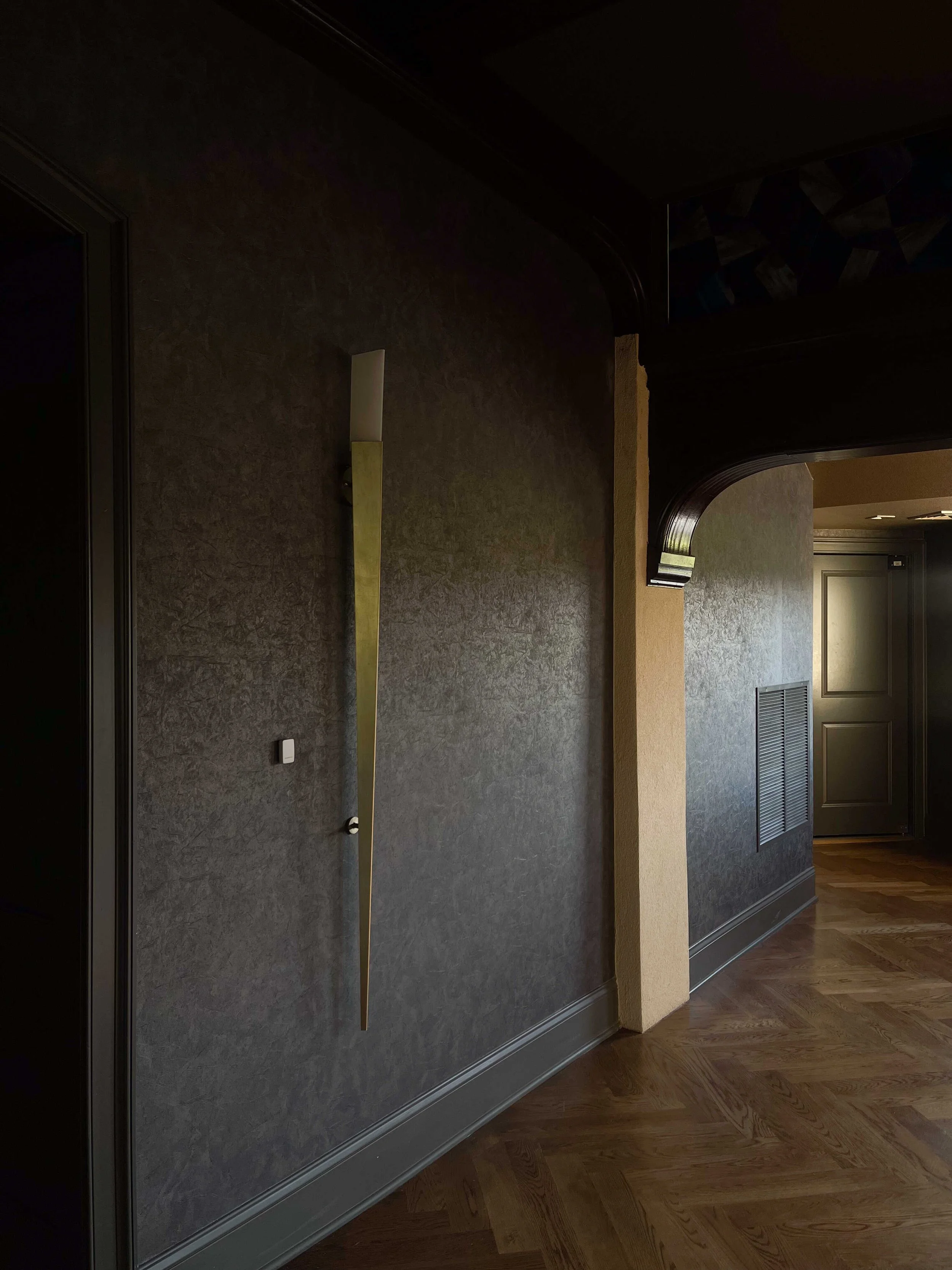 A moody, dark-themed hallway with textured charcoal wallpaper. A long, slender gold-tapered wall sconce provides a dramatic point of light against the dark walls and dark wood trim.