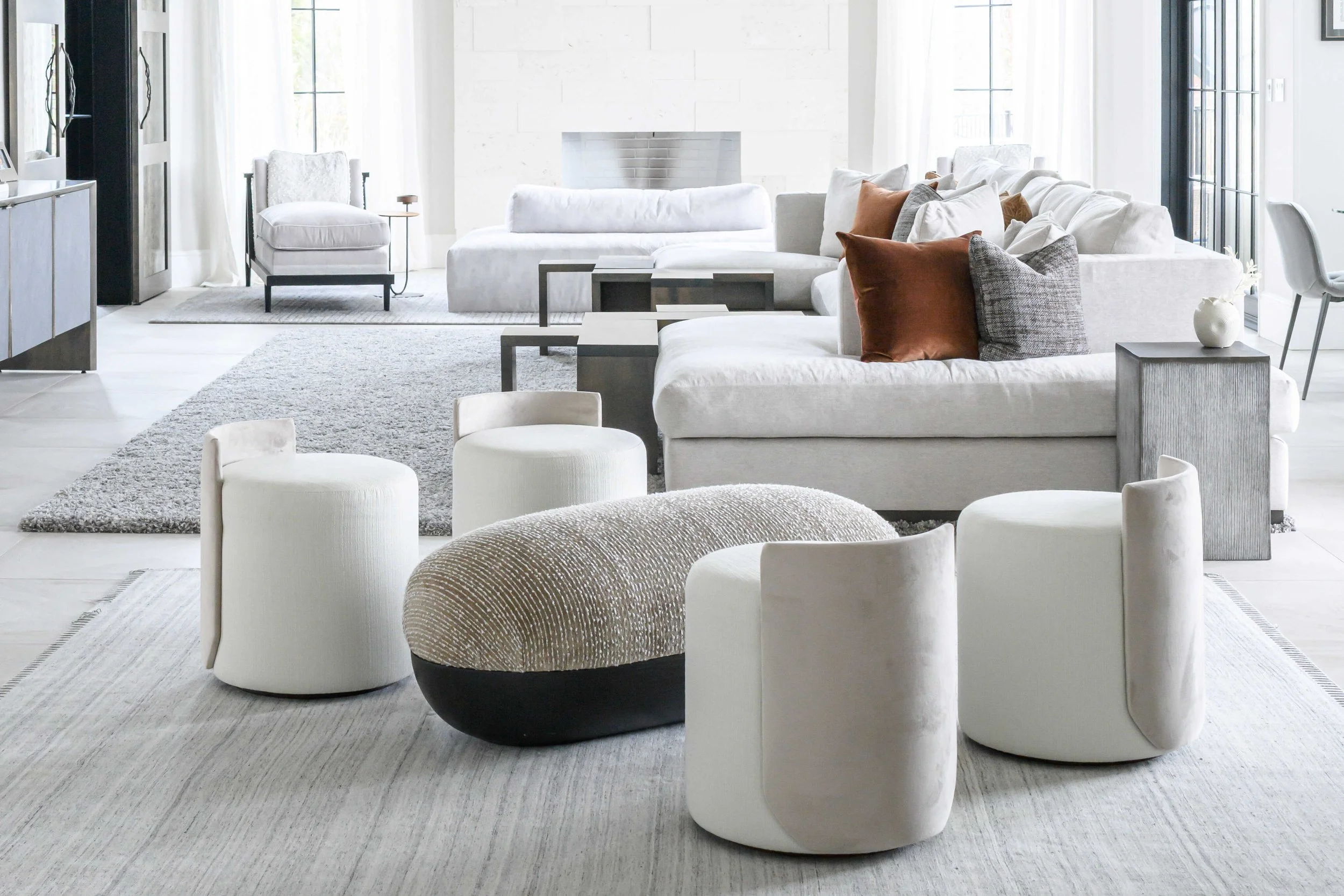Airy contemporary living room featuring white sofas with rust and gray accent pillows, cream cylindrical ottomans surrounding textured beige and black ottoman, nested modern coffee tables, and tall black-framed windows.