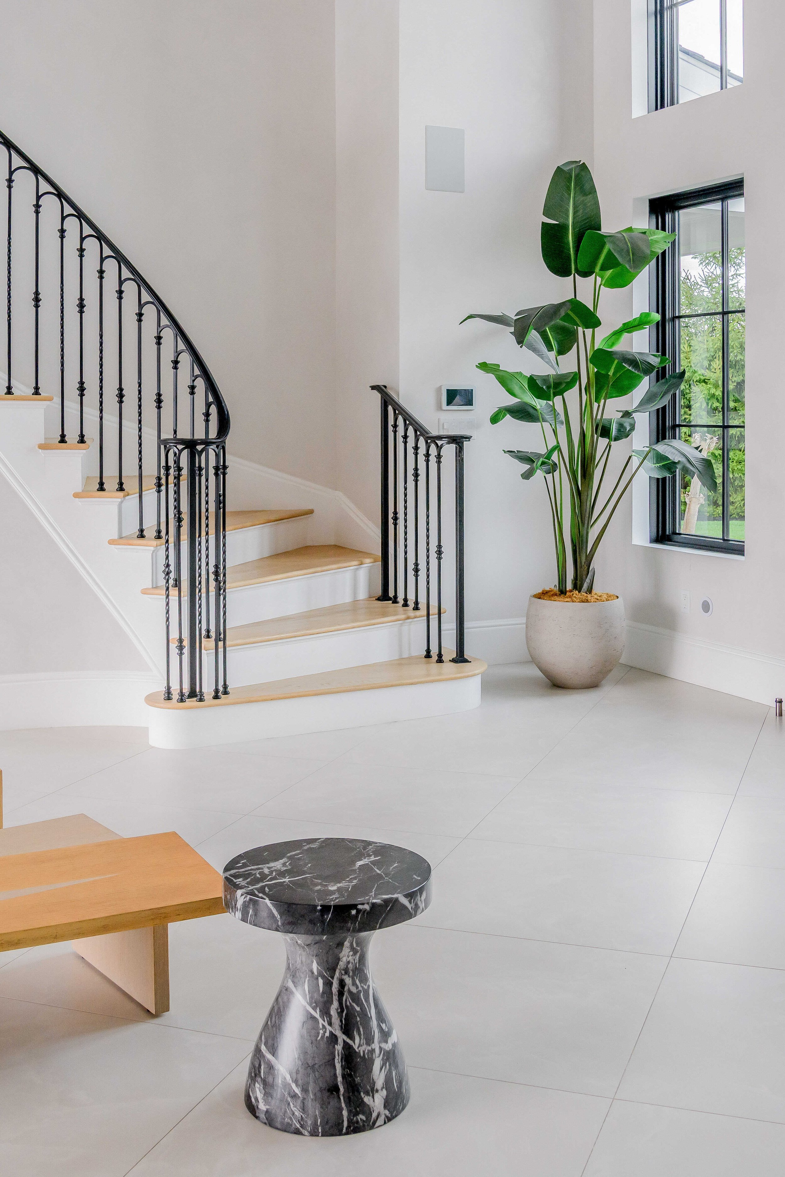 Bright contemporary staircase with light wood treads, white risers, and ornate black iron spindle railings, black-framed windows overlooking greenery, tall bird of paradise plant in cream planter, and black-and-white marbled pedestal side table in fo