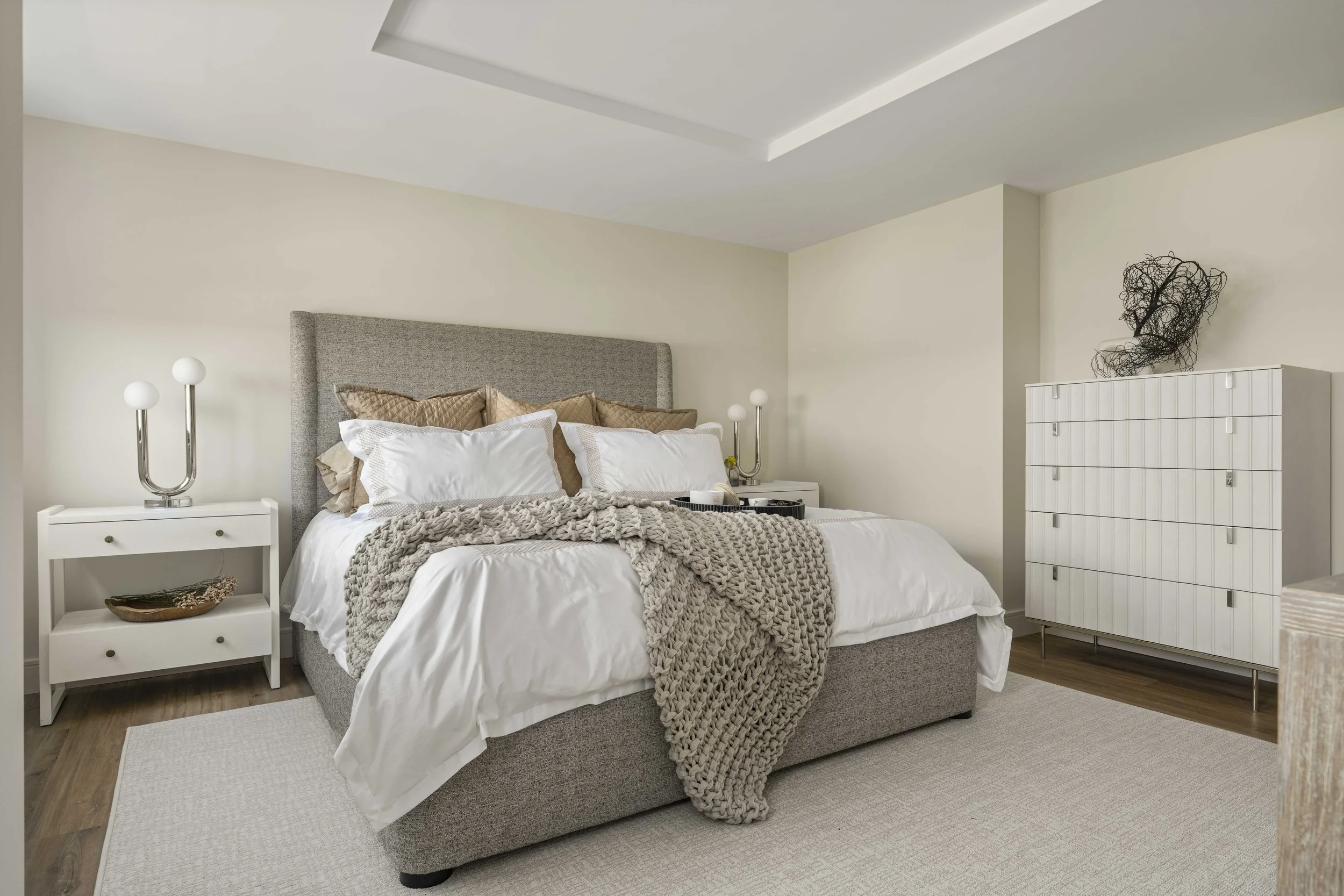 A bright, modern bedroom featuring a gray upholstered bed with white linens and a chunky knit gray throw. It is flanked by two white nightstands with unique U-shaped silver lamps, and a white dresser stands against the far wall.