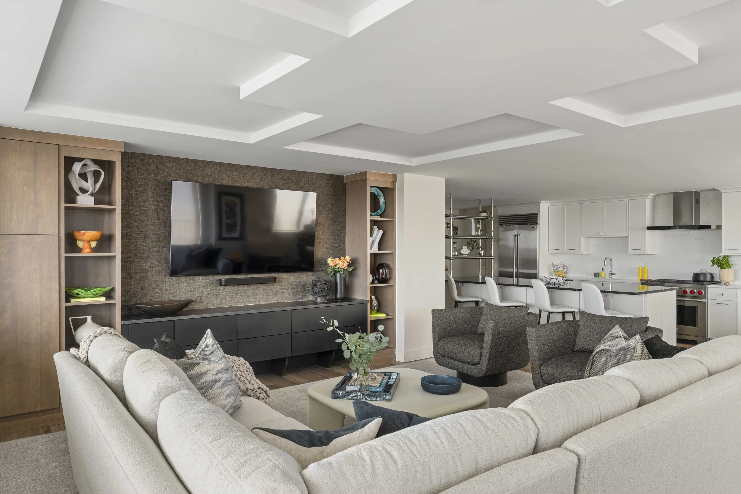 An open-concept living space showing a large cream sectional sofa, two gray swivel chairs, and a geometric coffered ceiling. The modern white kitchen is visible in the background.