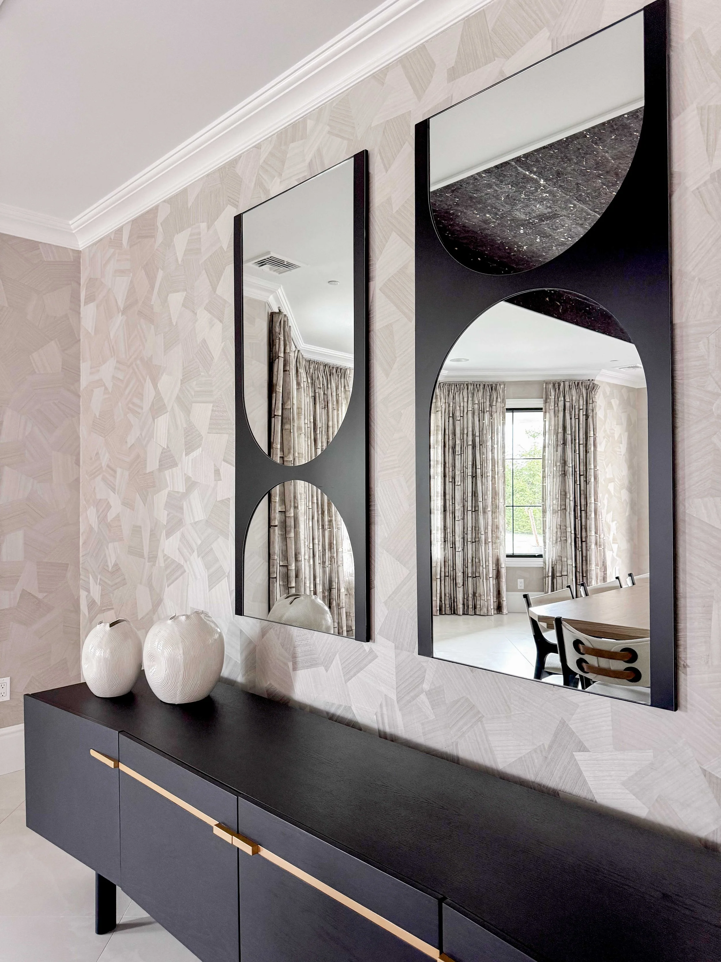 Elegant entryway vignette with floating black console featuring brass drawer pulls, pair of tall black-framed mirrors with curved geometric cutouts creating abstract shapes, white spherical ceramic vases, and textured neutral wallcovering.