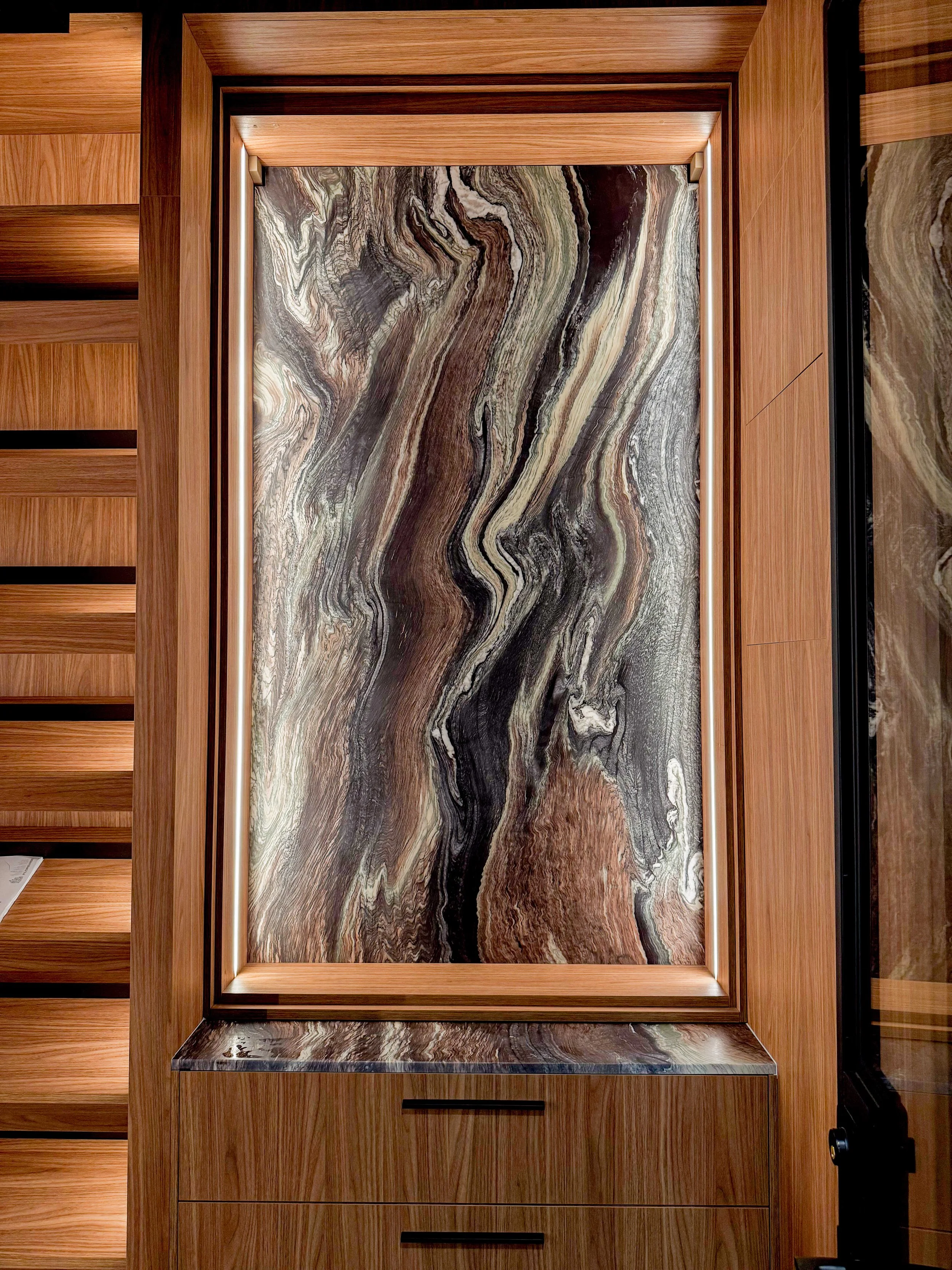 Illuminated display niche featuring dramatic swirled marble panel in browns, grays, and creams set within warm wood cabinetry with integrated LED lighting and matching marble shelf below.