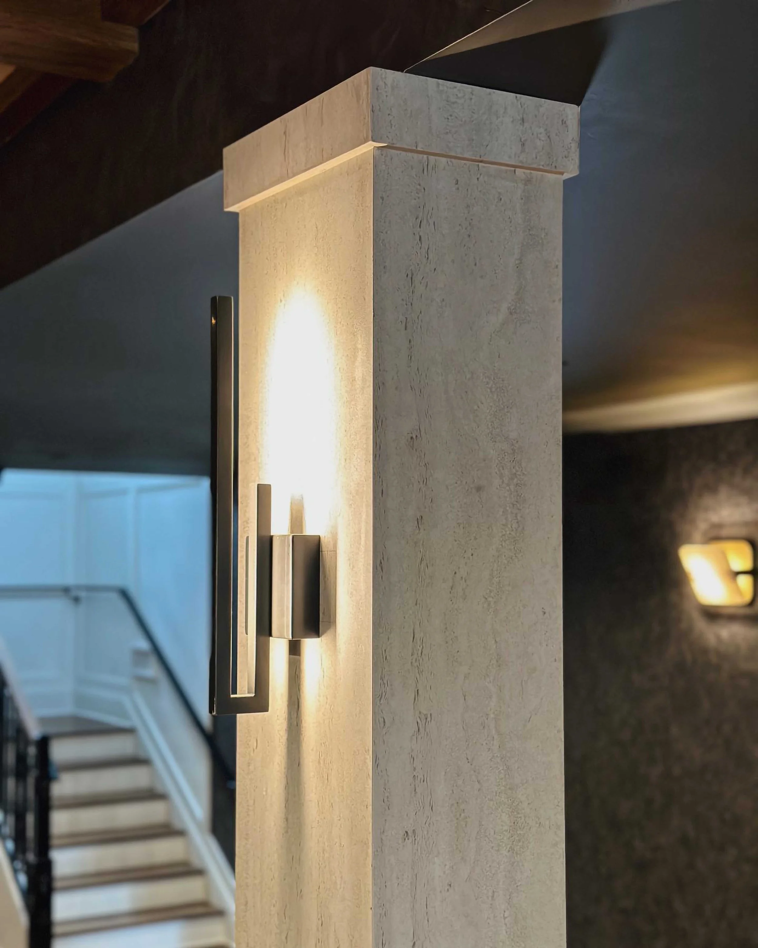 A close-up of a square architectural pillar with a textured cream finish, featuring a sleek, minimalist bronze wall sconce that casts a warm glow.