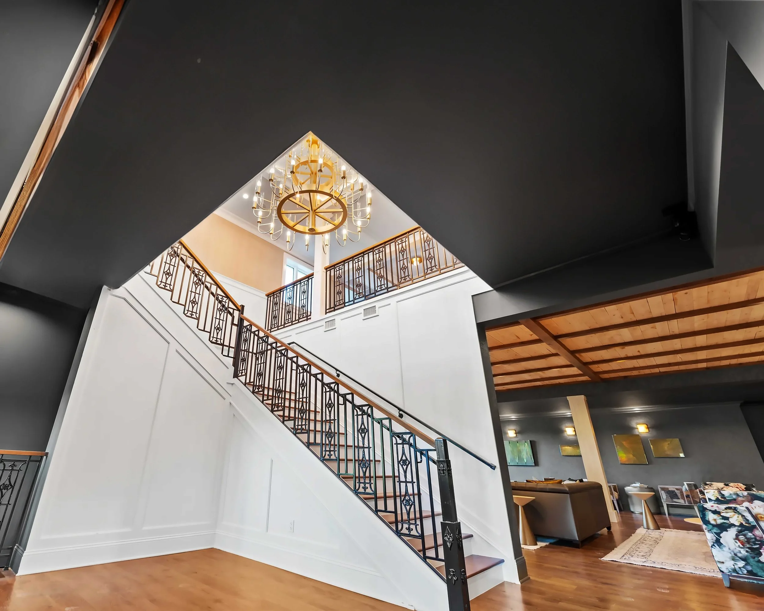 A wide-angle view of a grand staircase with white wainscoting and wrought-iron railings, illuminated by a large circular gold chandelier.