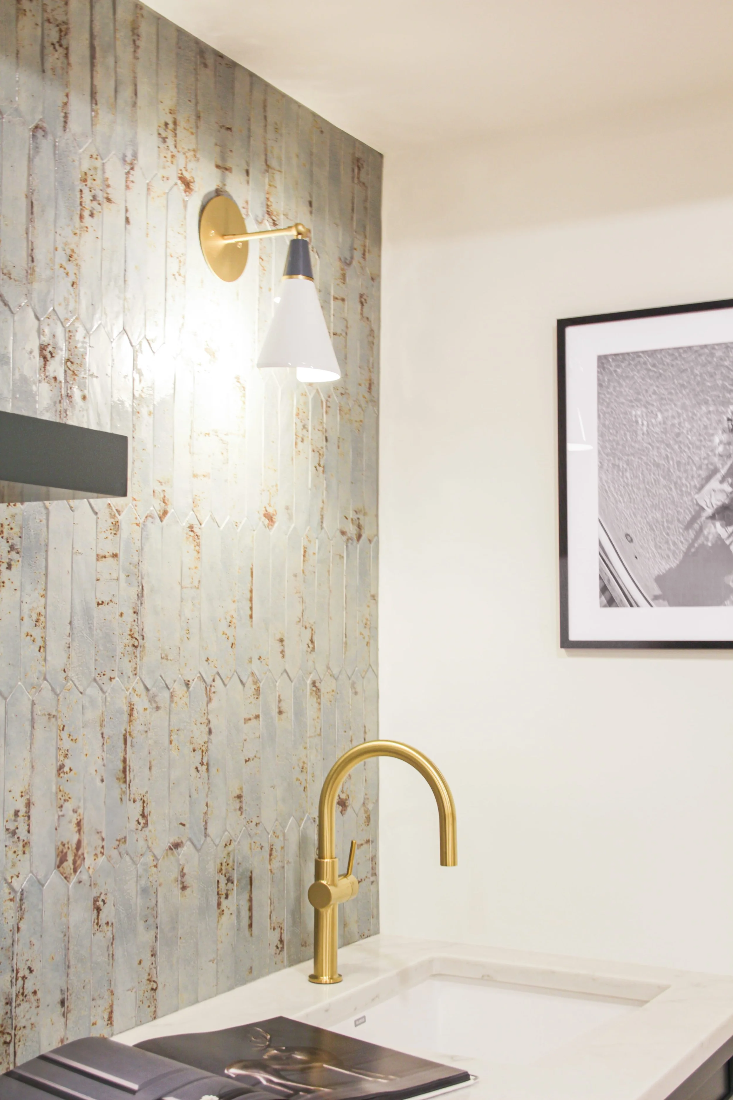 Detail of wet bar featuring brushed brass modern faucet on white countertop against distressed white patina tile backsplash, modern brass and white cone wall sconce, and black-framed artwork visible on adjacent wall.