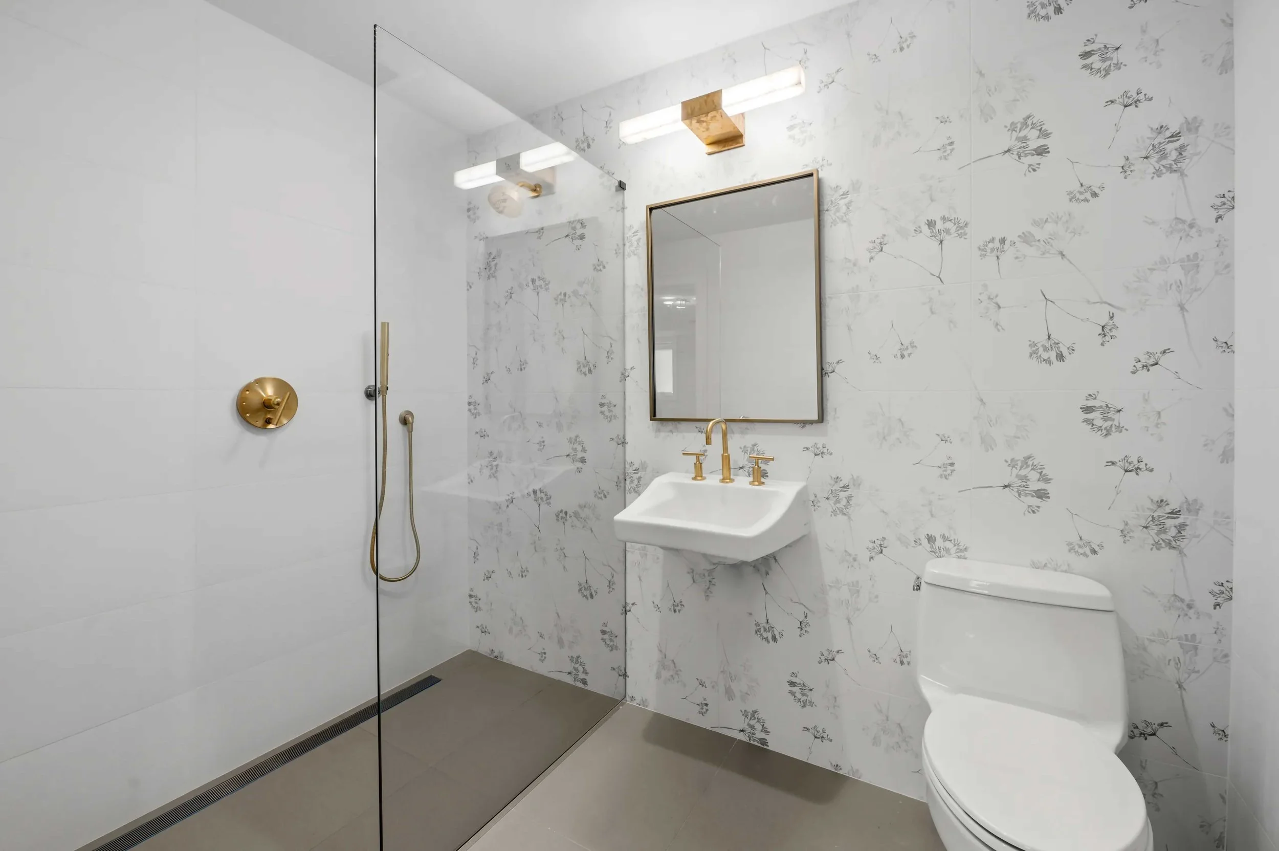 A clean, modern bathroom featuring a walk-in shower with a frameless glass partition and gold-toned fixtures. The wall behind the white pedestal sink and toilet is decorated with a delicate grey floral or botanical pattern. A rectangular mirror and a