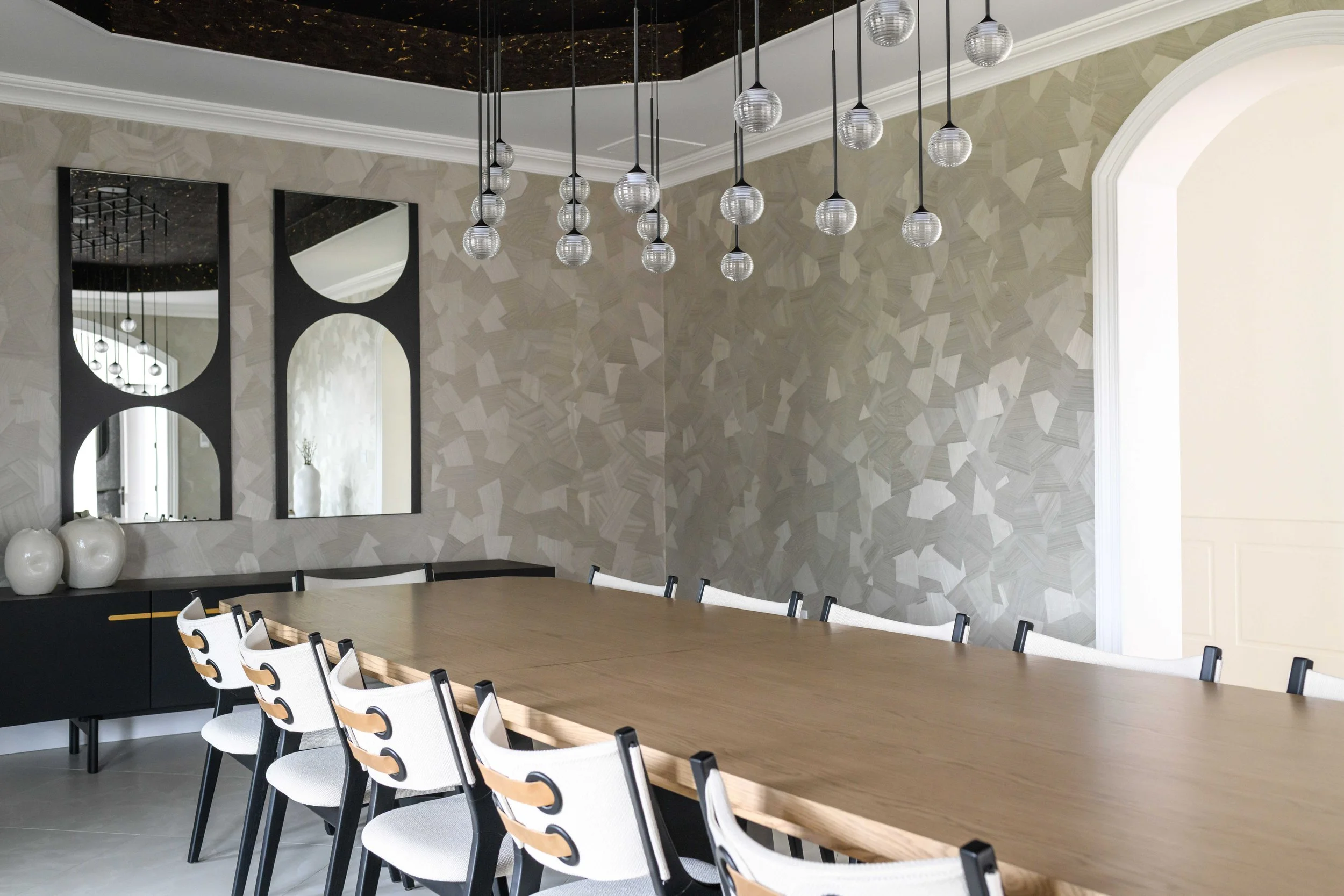 Elegant dining room with oval wood table, white chairs with black frames and circular cutout details, cluster of glass globe pendant lights, textured neutral geometric wallcovering, and black ceiling detail.