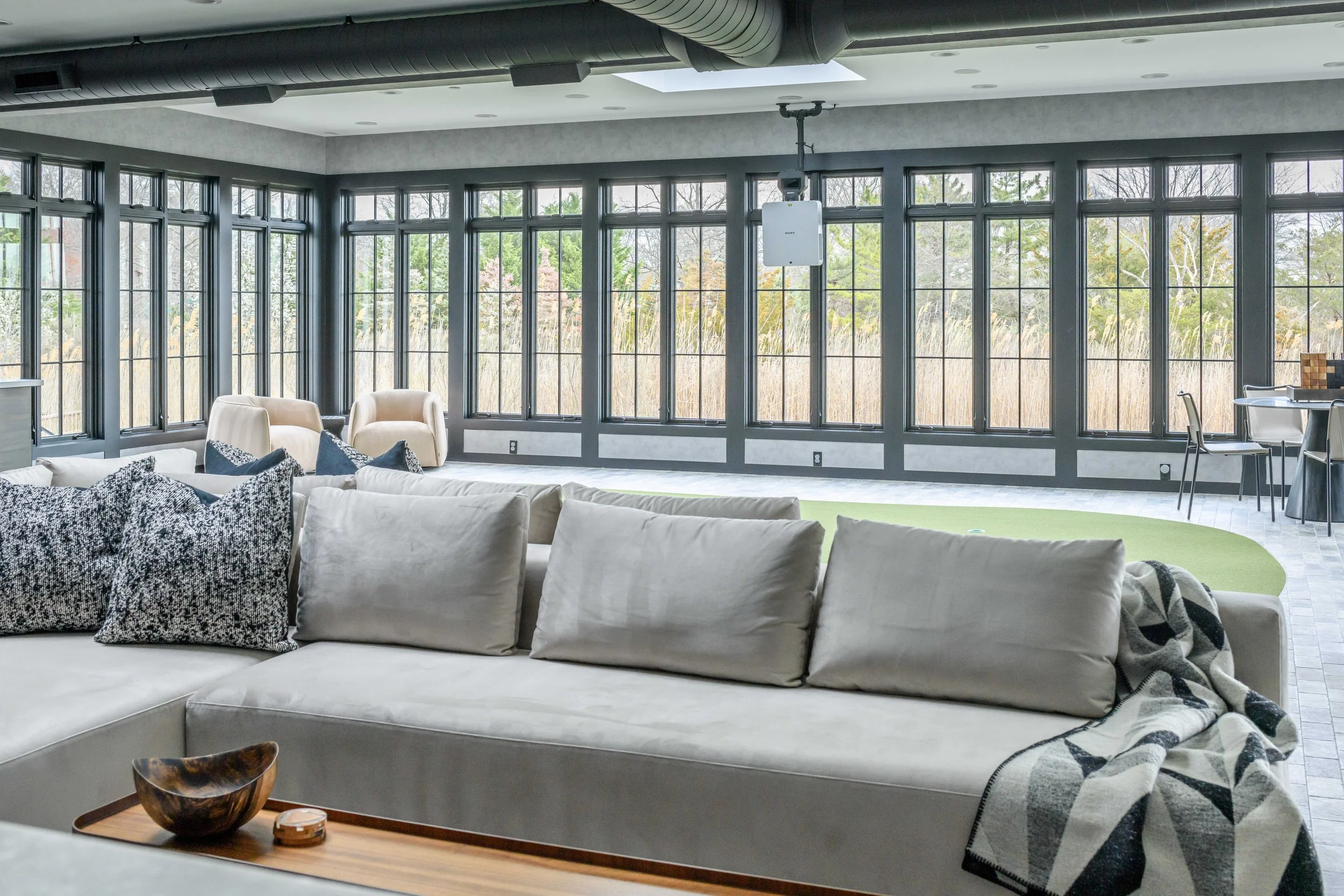 Bright sunroom with wall of black steel-framed windows overlooking green space, low gray sectional with patterned throw pillows, and cream upholstered seating visible in background.