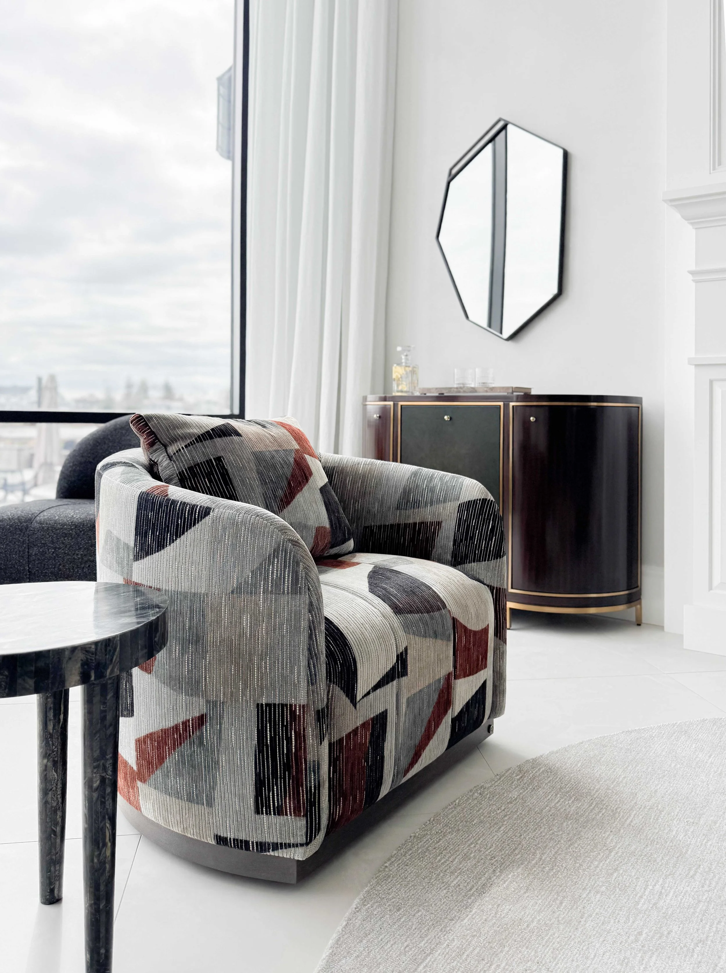 Detail shot of modern swivel chair upholstered in abstract geometric fabric with gray, black, and rust tones, hexagonal black-framed mirror on wall, dark wood credenza with brass trim, and floor-to-ceiling windows.